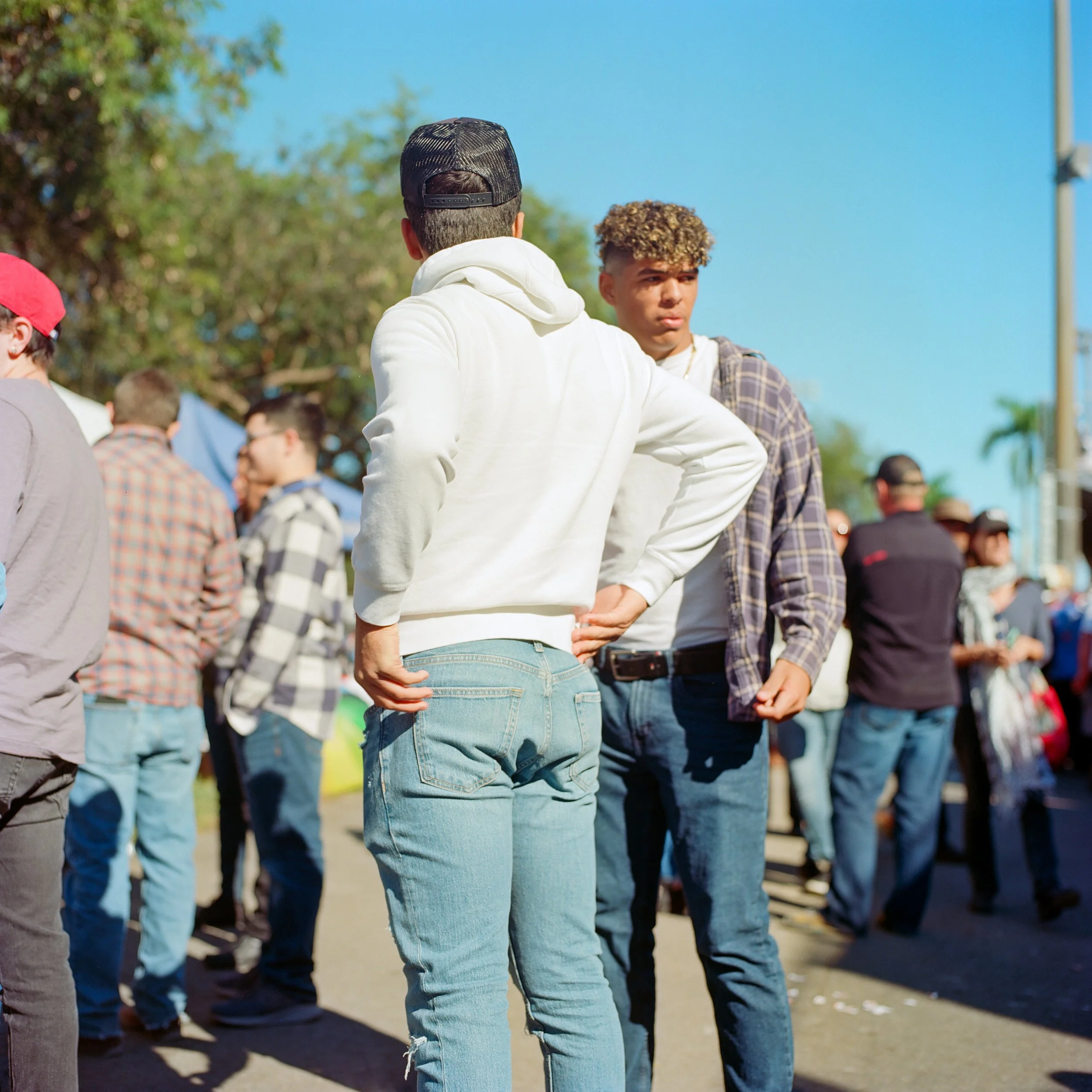 People standing outdoors at a daytime event, some chatting, with a clear blue sky and trees in the background.