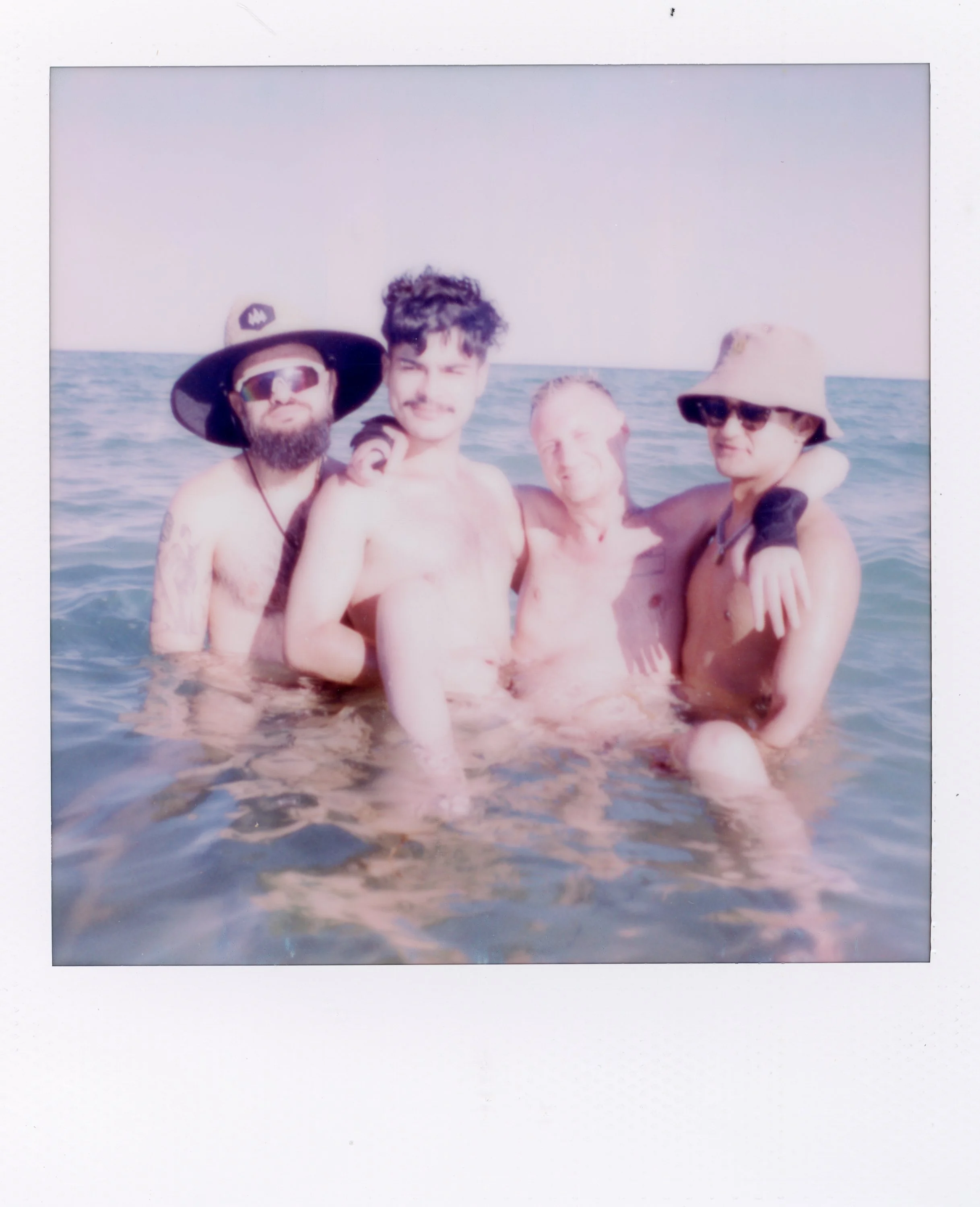 Four young men are standing in the ocean, shirtless, with one wearing a large sun hat and sunglasses, and another wearing a baseball cap and sunglasses. They appear happy and are closely grouped, with some arms around each other, enjoying a sunny day