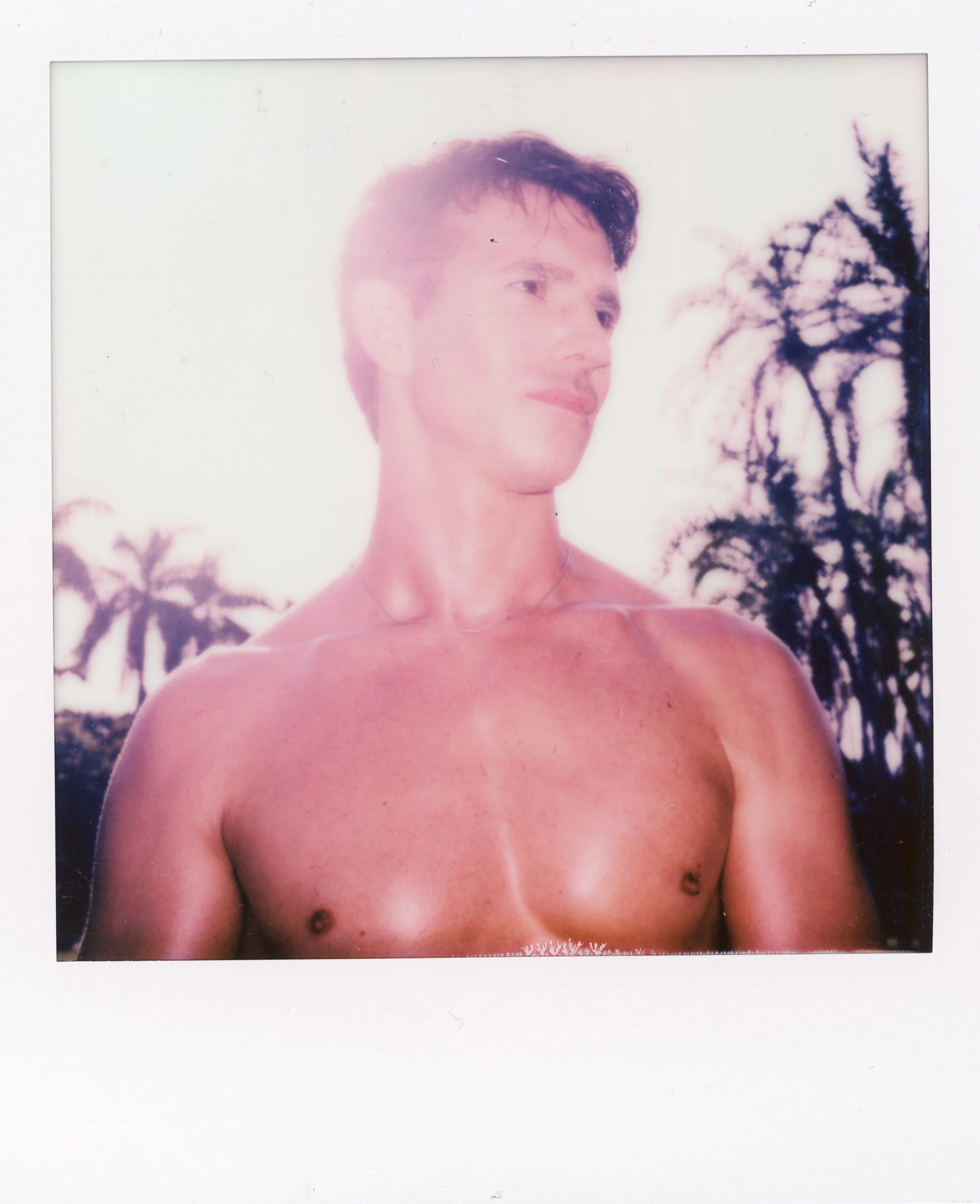 A shirtless young man with short dark hair and a necklace, standing outdoors with palm trees in the background, under a bright sky.