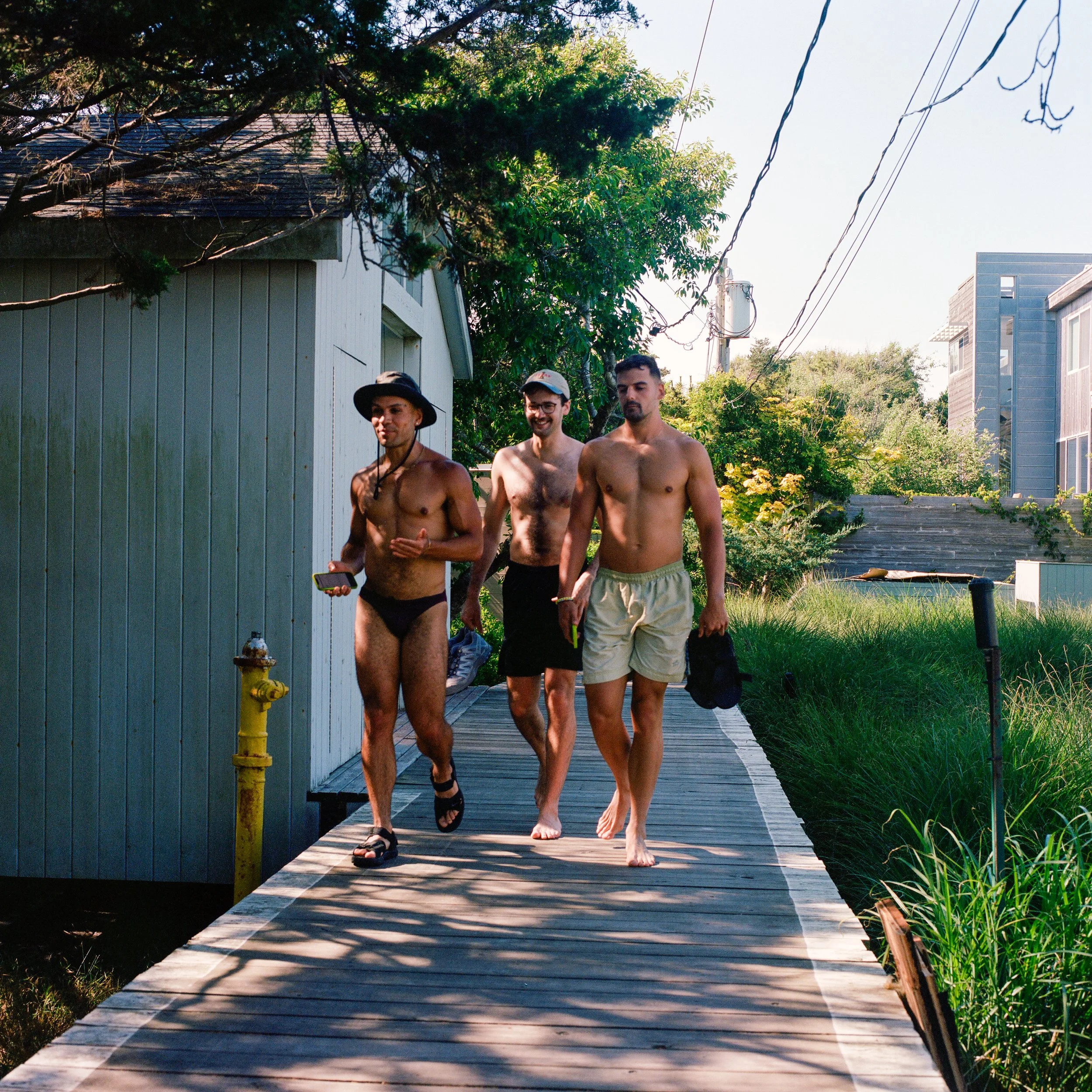 Three shirtless men walk barefoot on a wooden boardwalk in a garden area. One man wears black shorts, a black hat, and sandals, holding a phone. The second man wears black shorts and glasses, carrying shoes. The third man wears beige shorts, holding