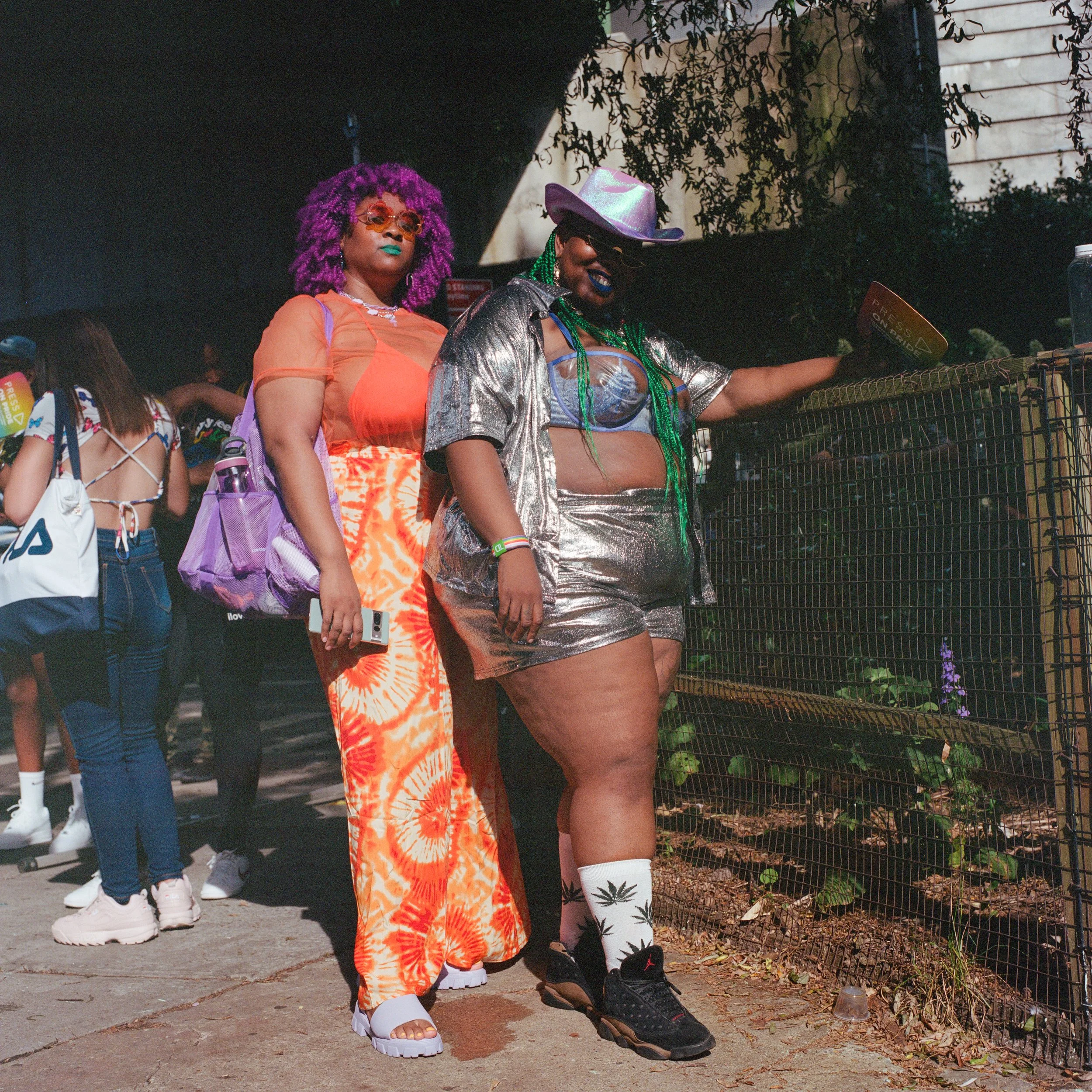 Two women standing in a sunlit outdoor area at a pride event. One woman has purple curly hair, sunglasses, and bright makeup, wearing orange tie-dye pants and a sheer orange top. The other woman has green braids, wearing a metallic silver outfit, a p