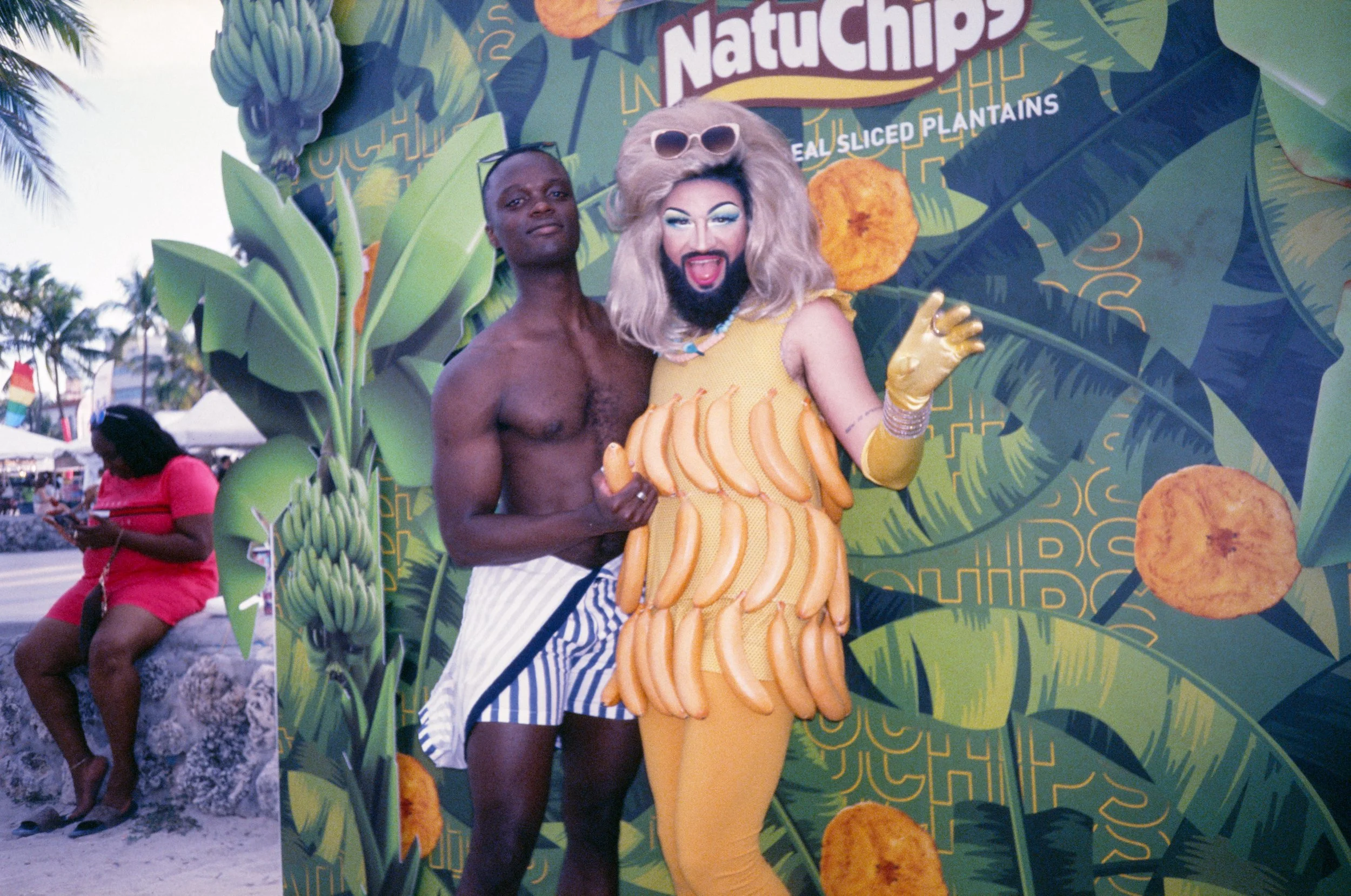 A shirtless man in striped shorts standing next to a person dressed in a banana-themed costume with sunglasses on their head, standing in front of a Natuchips advertisement with banana leaves and potato chips. In the background, a woman in a pink dre