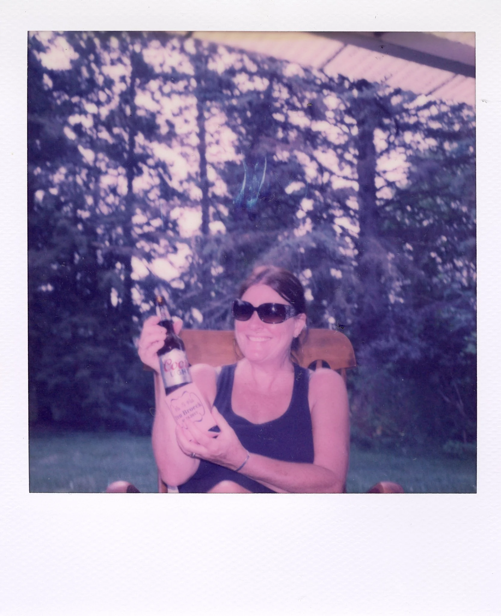 Woman sitting outdoors in a wooden chair, wearing sunglasses, smiling, holding a bottle of Coors Light beer, with trees in the background.