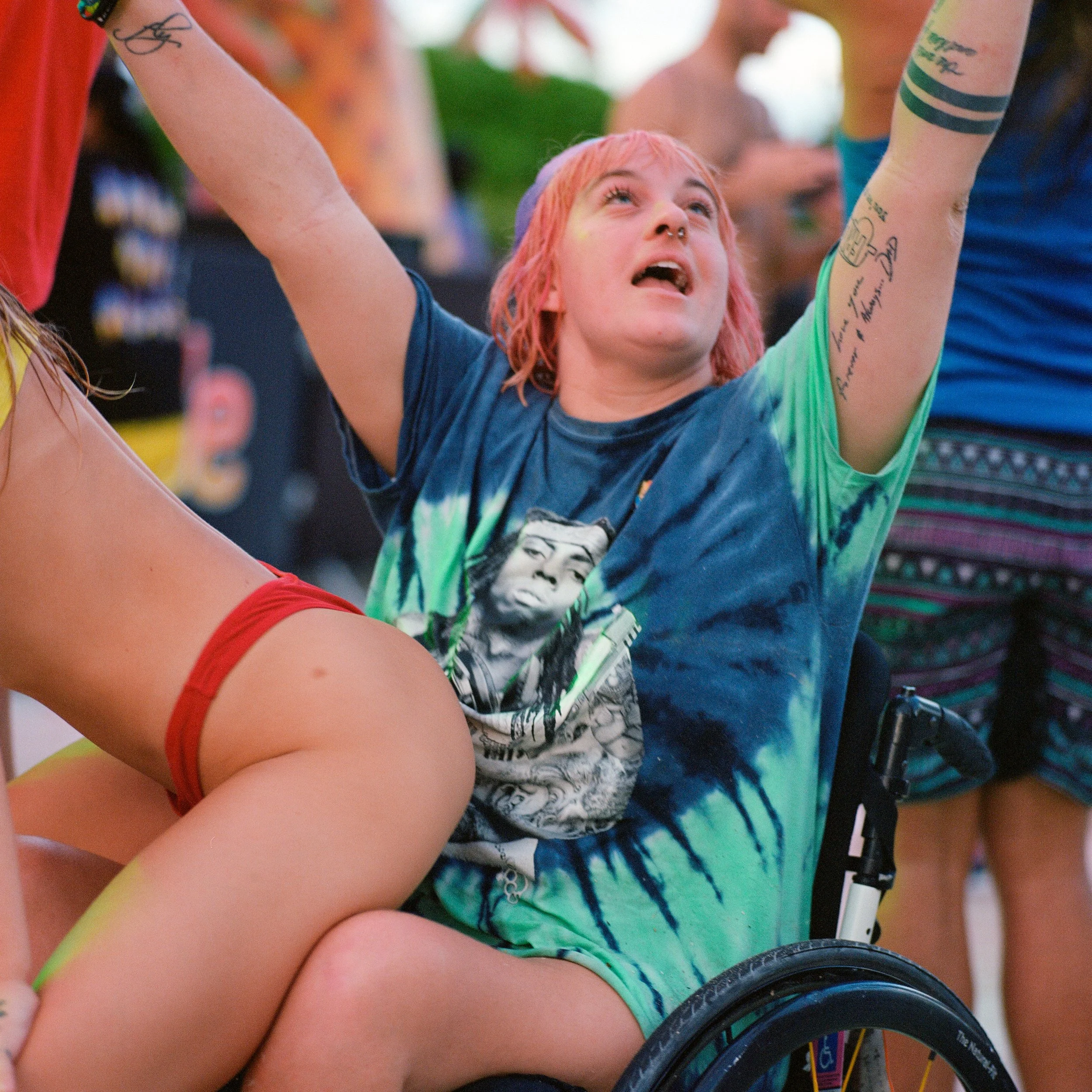 Person in a wheelchair with pink hair raising their arms, surrounded by other people, at an outdoor event.