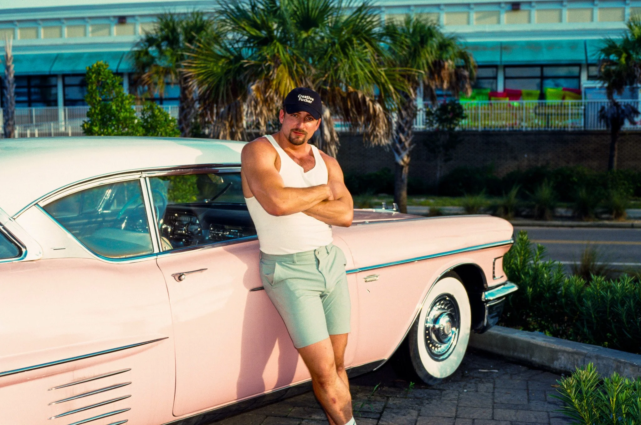 A man with crossed arms wearing a white tank top, light green shorts, a black cap, and white socks standing next to a vintage pink and white car with palm trees and a colorful building in the background