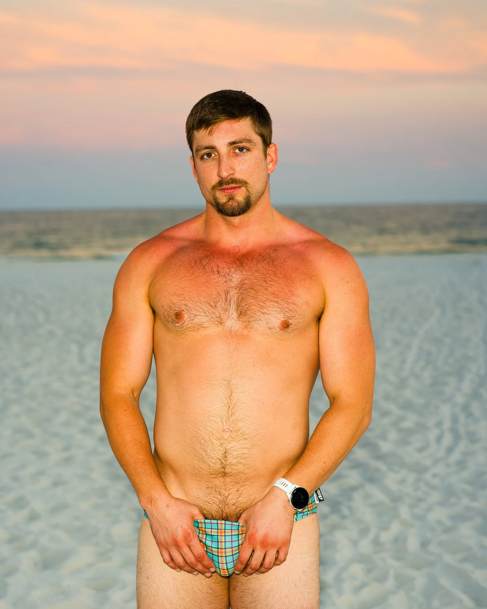A man standing on a beach at sunset, wearing colorful plaid swim briefs and a smartwatch.