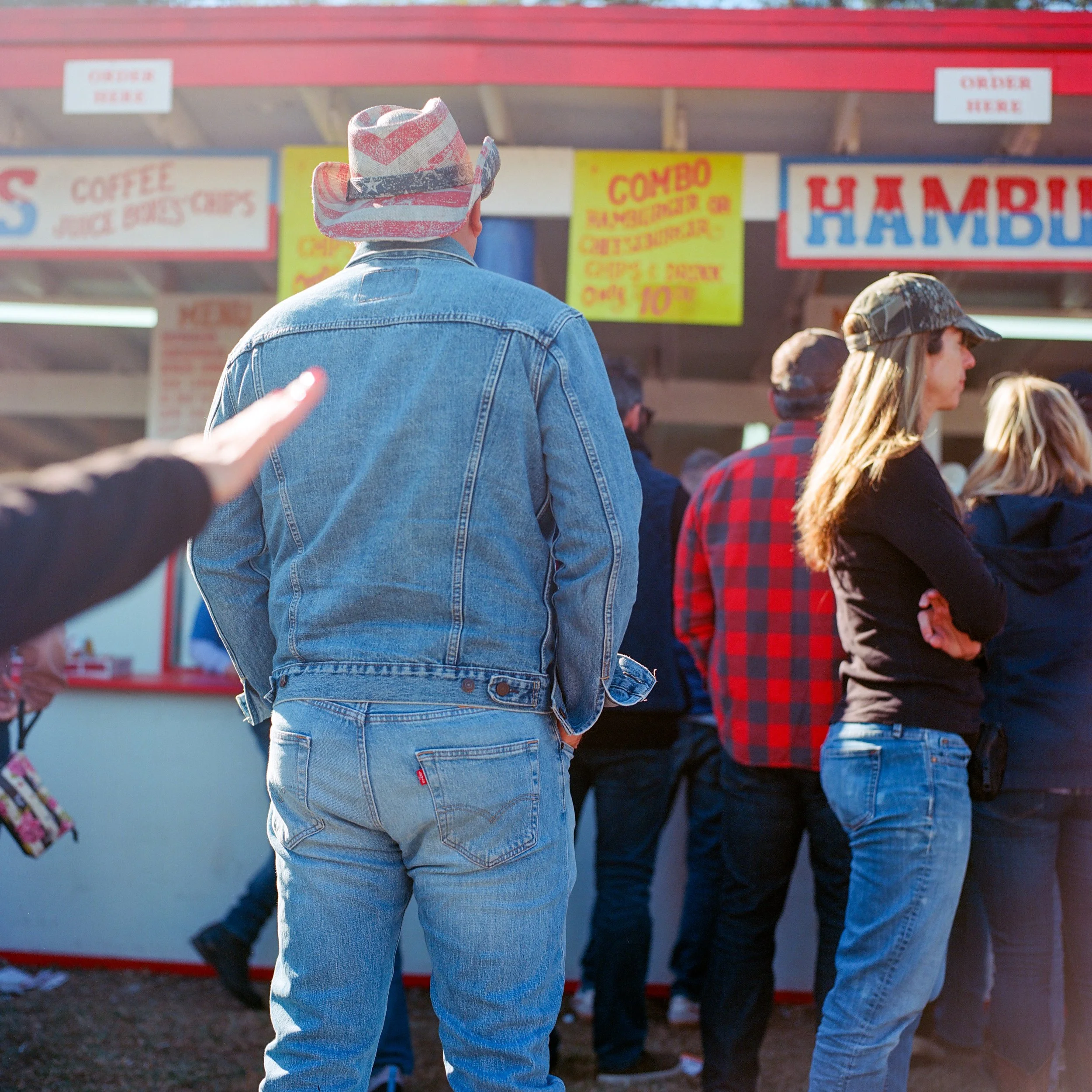 A man wearing a denim jacket and jeans with a patriotic star-spangled hat stands in line at a food booth at a fair, with other people waiting nearby.