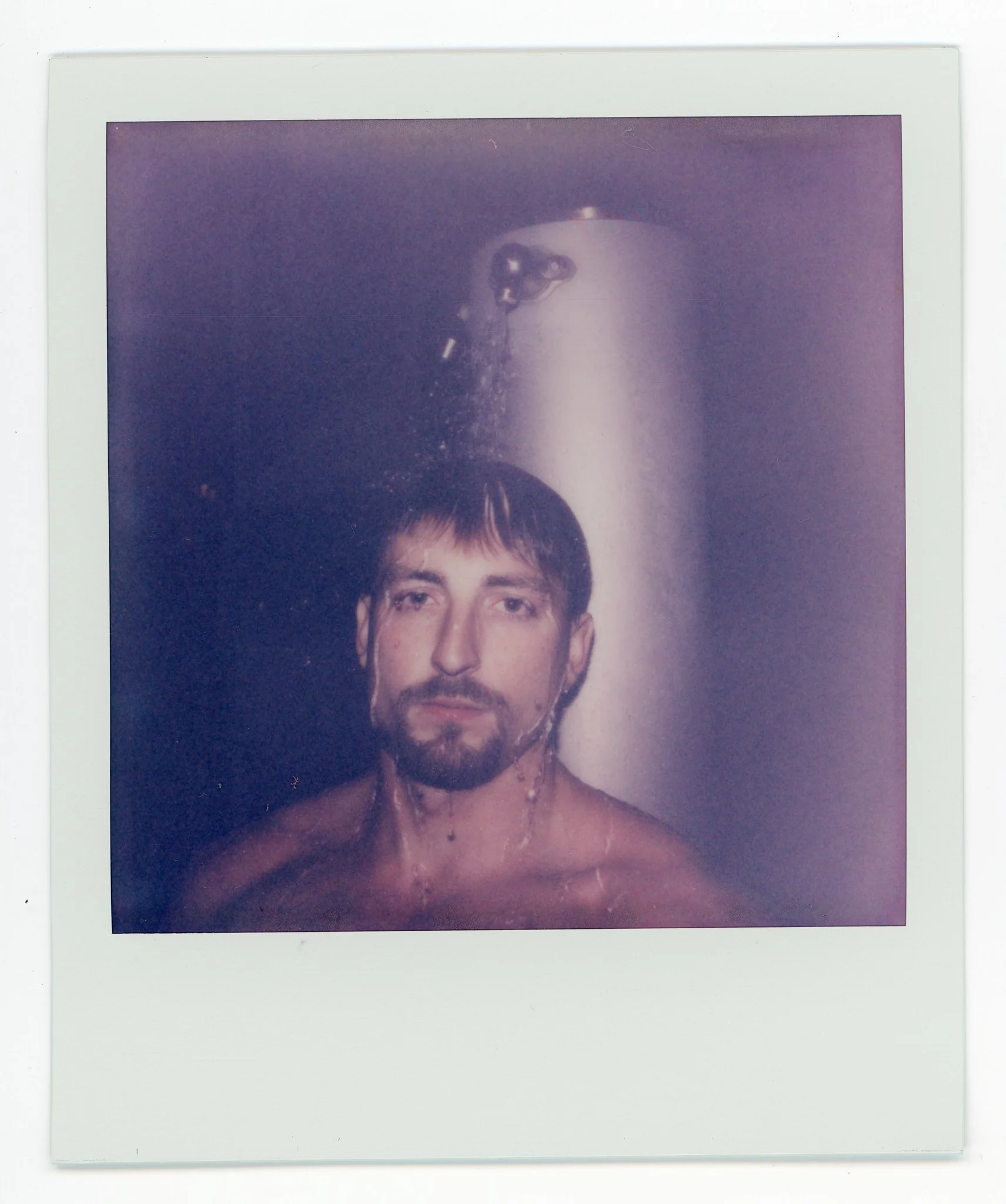 A man with short brown hair and a beard under a stream of water in front of a metallic cylindrical surface.