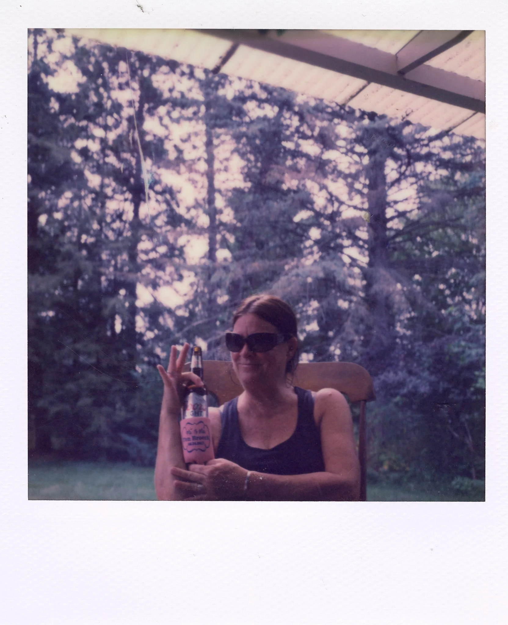 A woman wearing sunglasses sitting on a wooden chair outdoors, holding a beverage with a printed label, with trees and a canopy in the background.