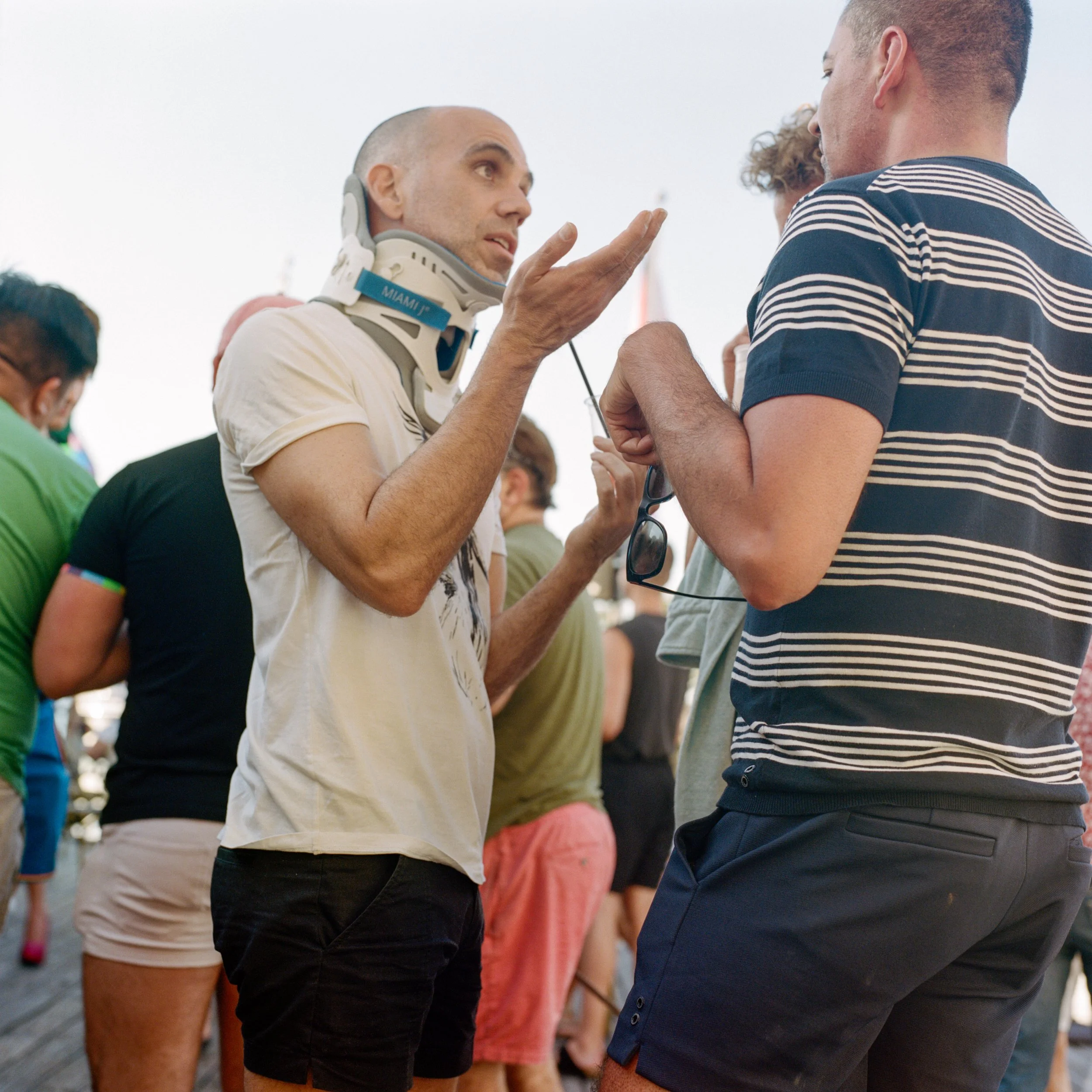Two men engaged in conversation among a group of people outdoors, one of whom is wearing a neck brace and gesturing with his hand.