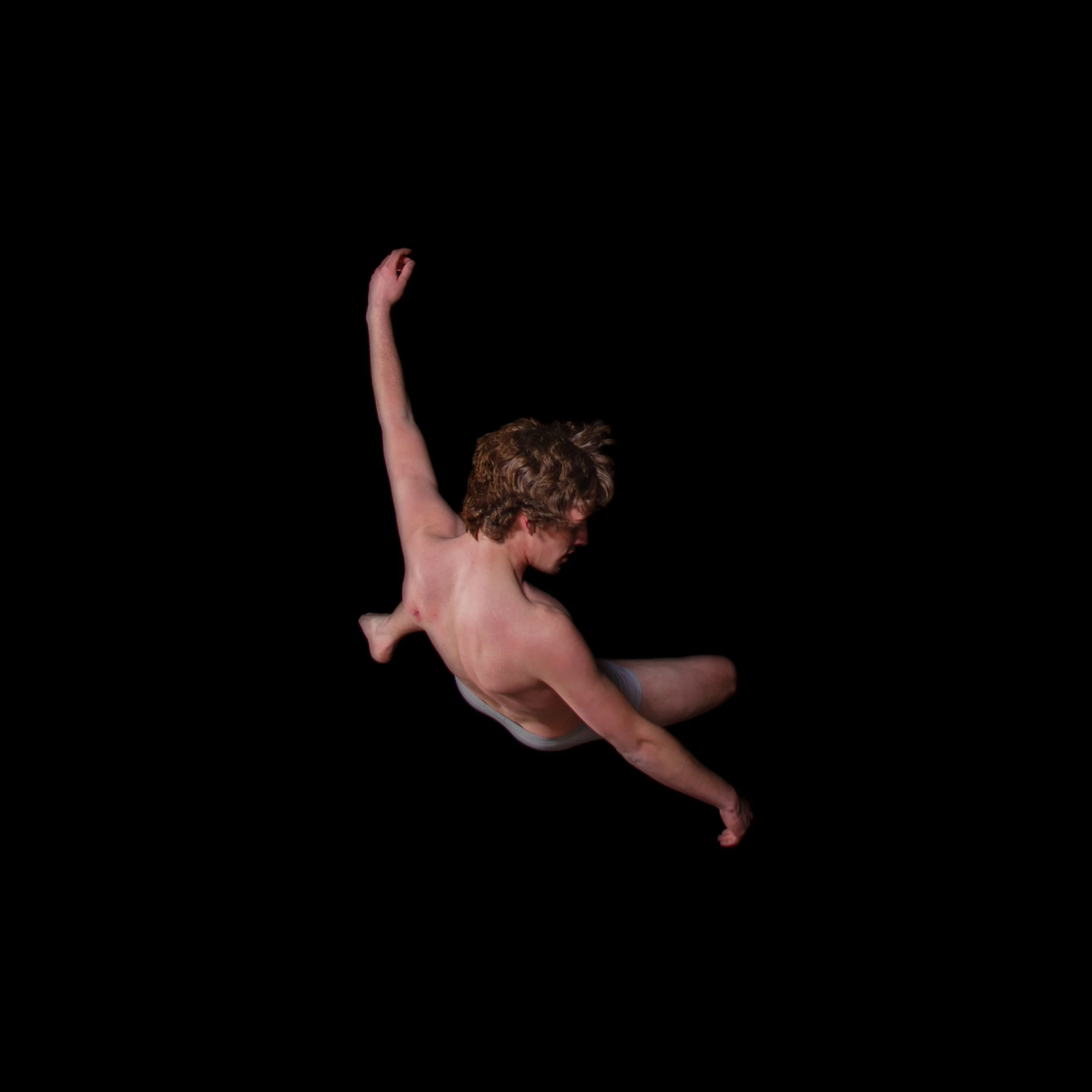 A young man with curly hair in mid-air, performing a dance move against a black background. He is shirtless and wearing gray shorts.