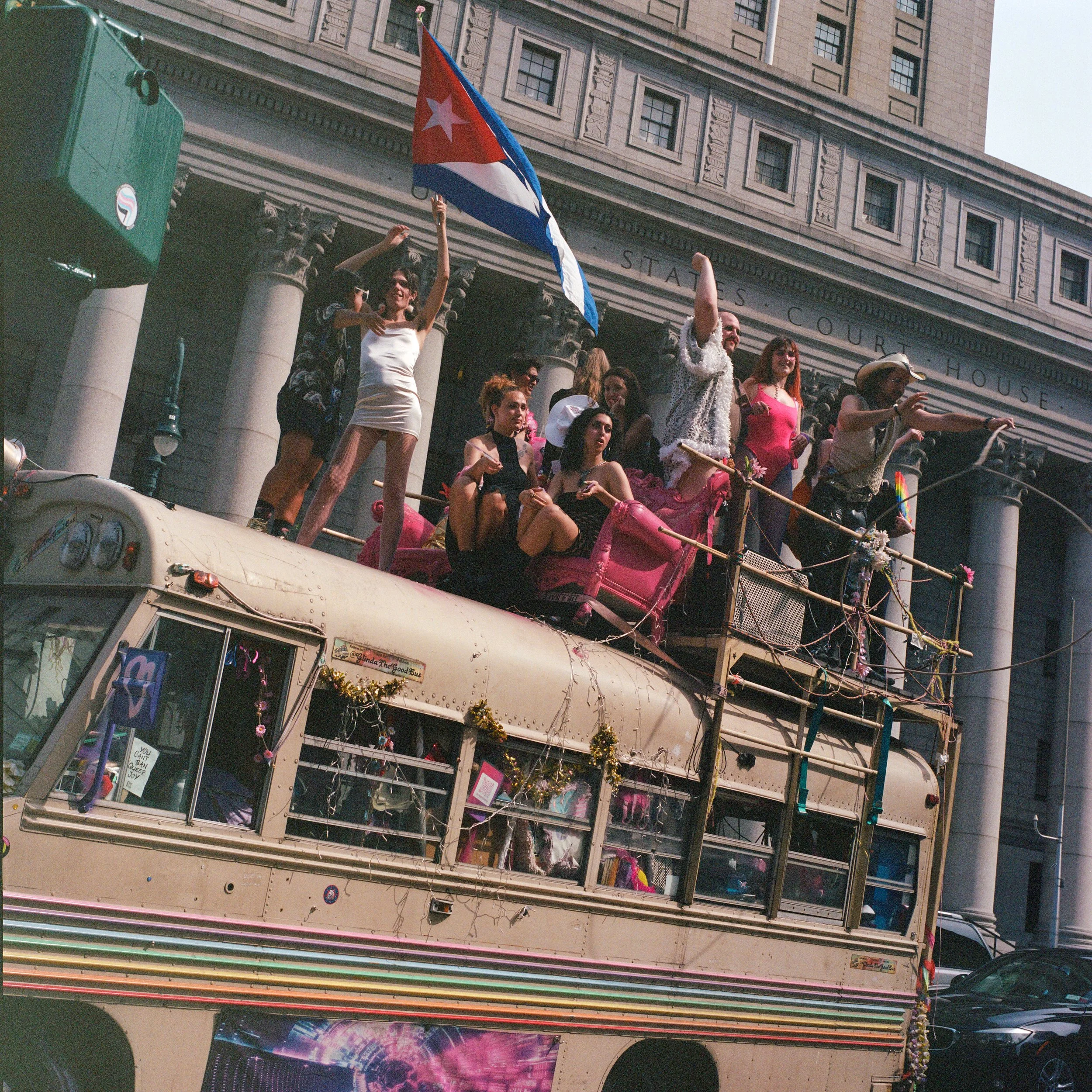People celebrating on a decorated double-decker bus during a pride parade, with a building labeled 'Courthouse' in the background.