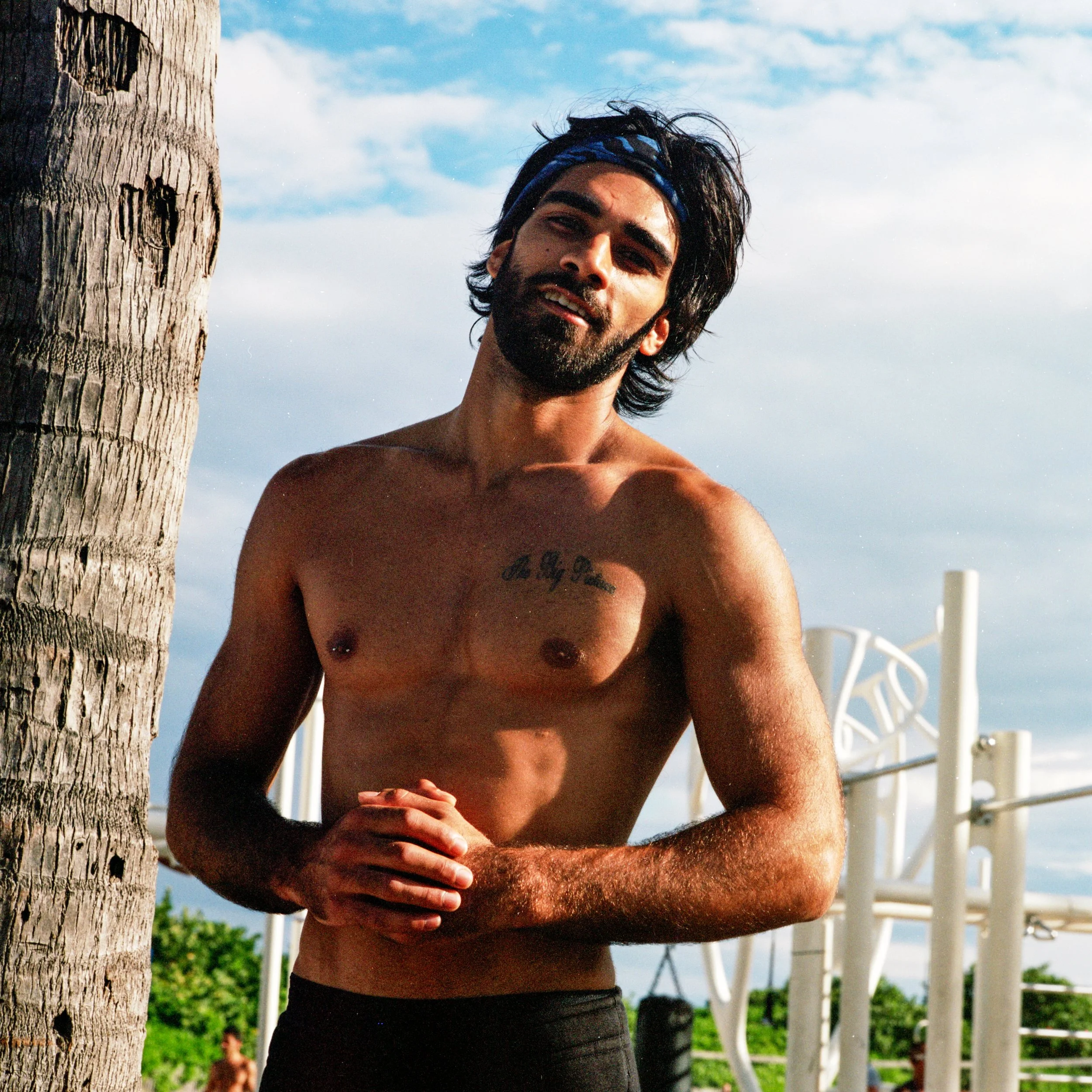 A shirtless man standing outdoors near a palm tree, with a blue bandana on his head, smiling at the camera, with sky, greenery, outdoor gym equipment, and blue sky in the background.