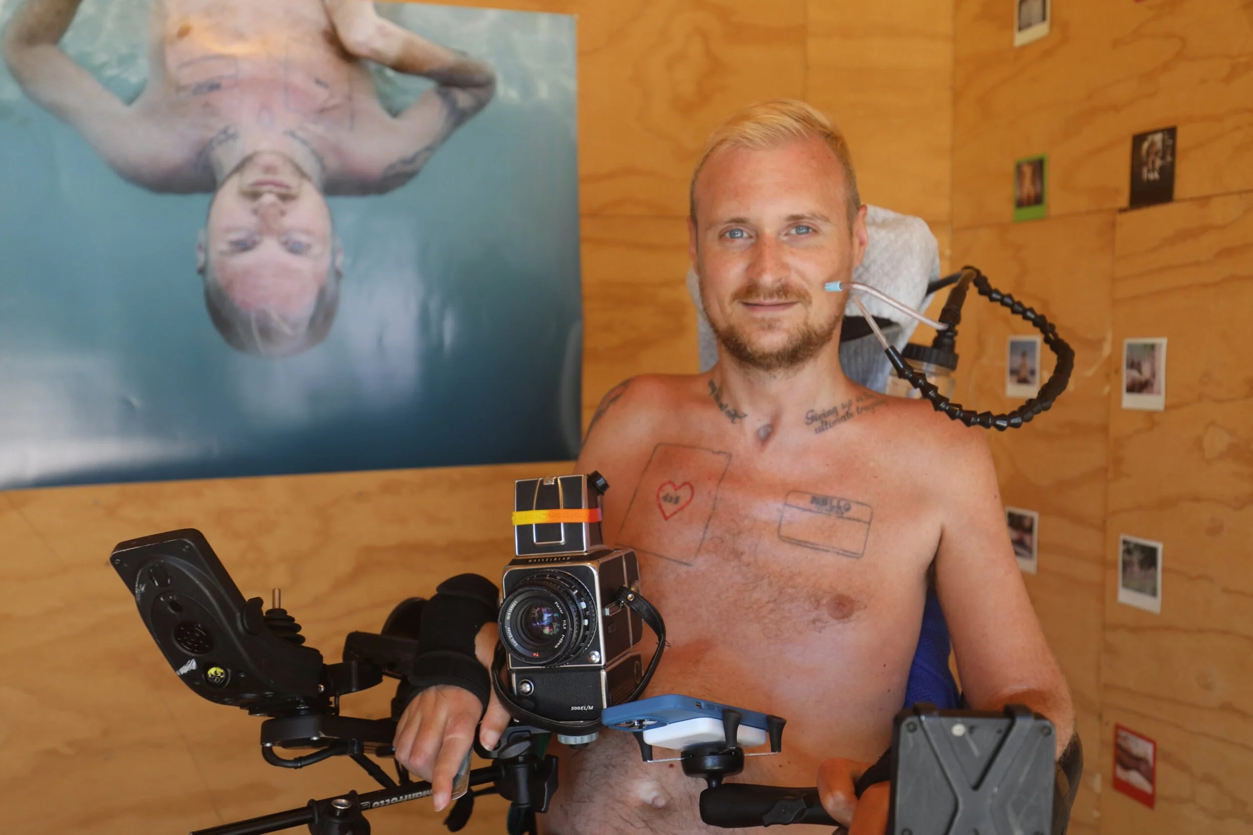 A man with a wheelchair, tattoos, and wearing no shirt, sitting in front of a wooden wall with photos, smiling, with a large photo of himself swimming behind him.