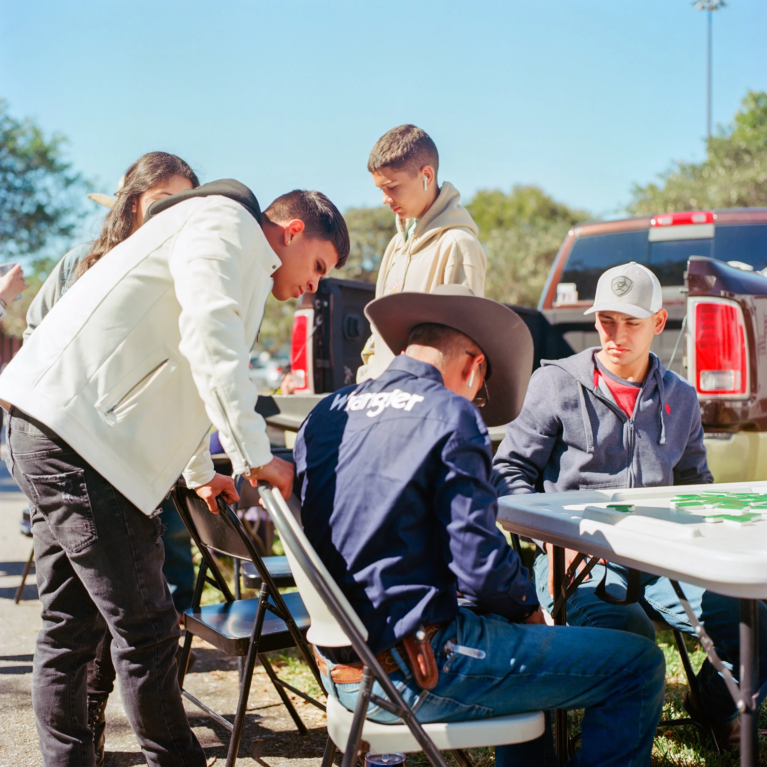 Group of young people gathered around a table outdoors, playing a game or studying, with backpacks and a pickup truck in the background on a sunny day.