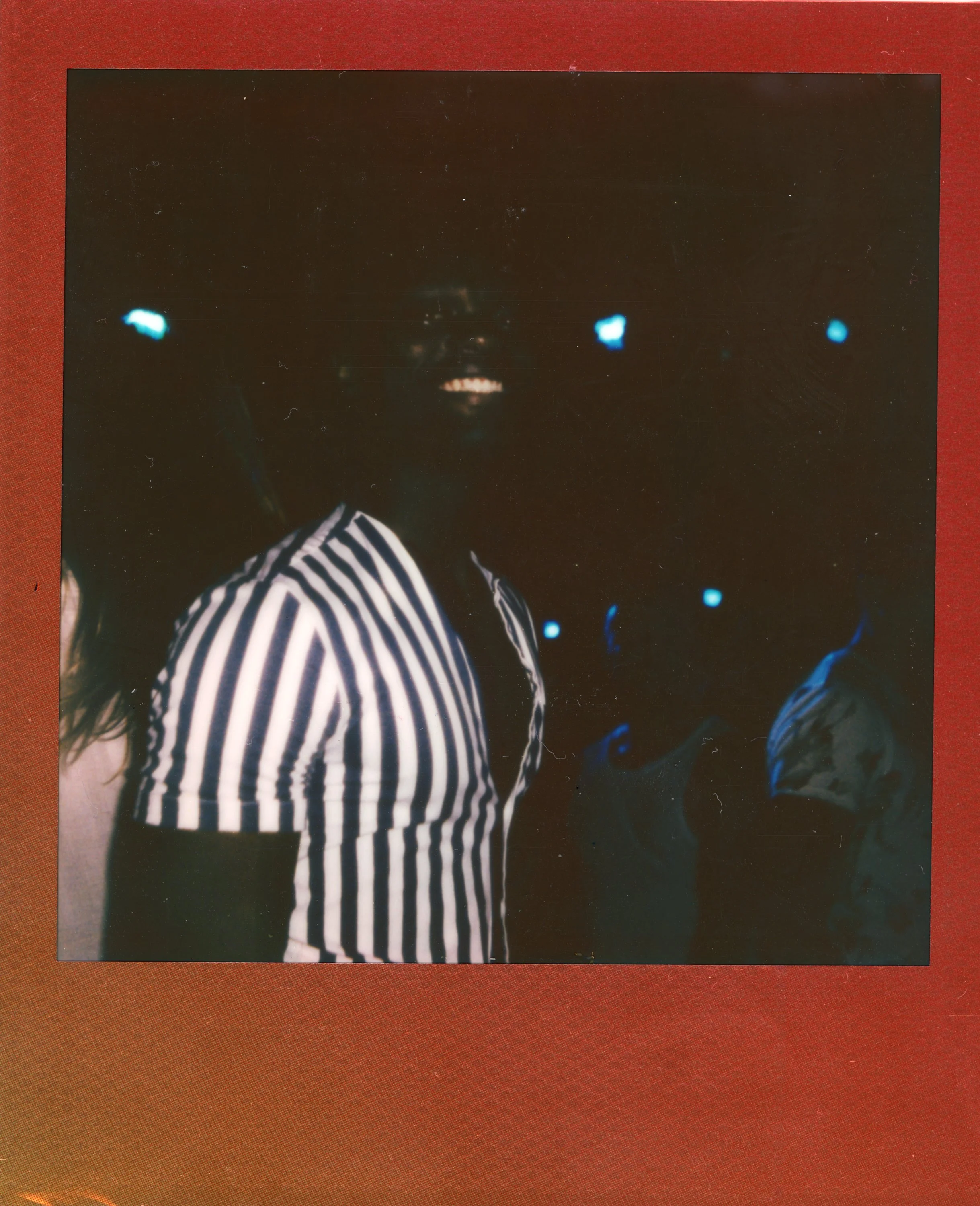 A person with a broad smile wearing a black and white striped shirt at a dimly lit event or party, surrounded by other people with blue-tinted lights in the background.