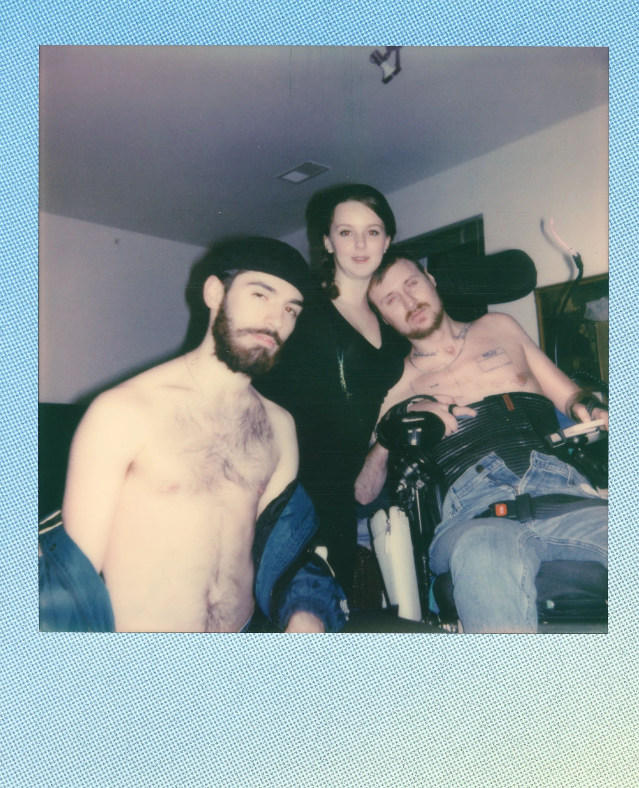 Three young adults indoors, two men without shirts, one woman in black, posing together, with one man in a wheelchair.