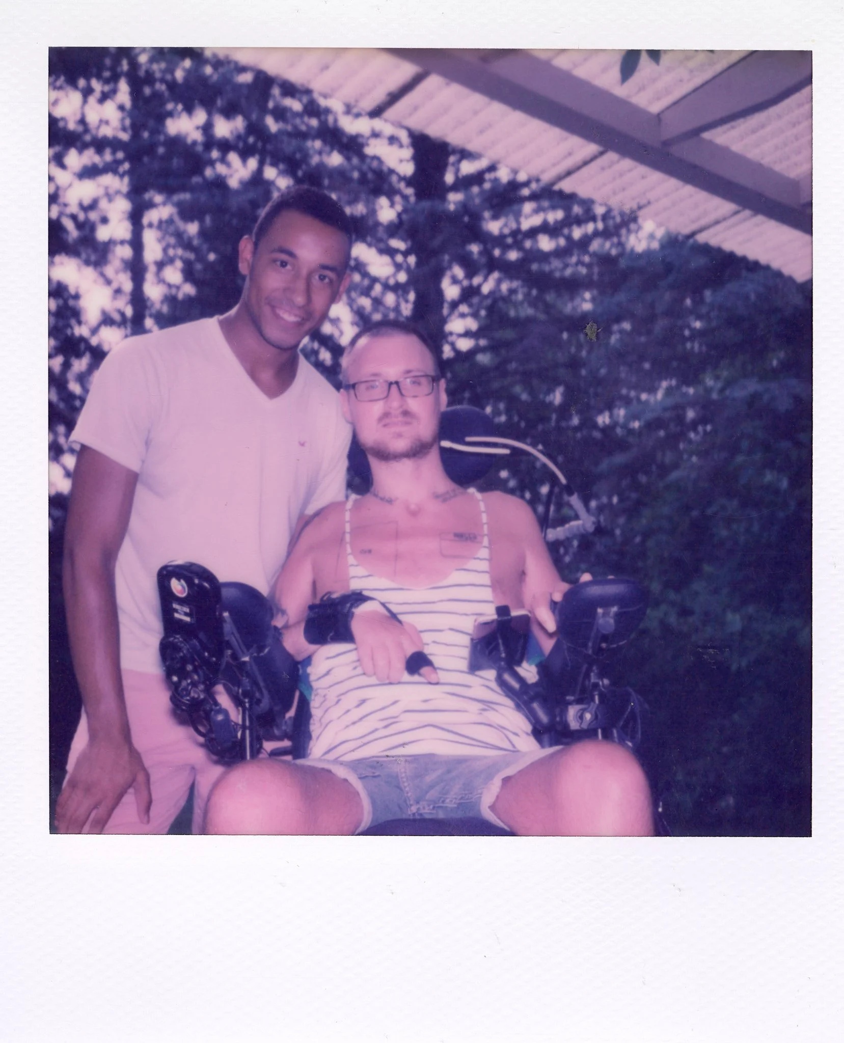 Two men, one standing and one seated in a motorized wheelchair outdoors under a metal roof, with trees in the background. The man in the wheelchair is wearing a striped tank top and denim shorts. The other man is wearing a white t-shirt and pink shor