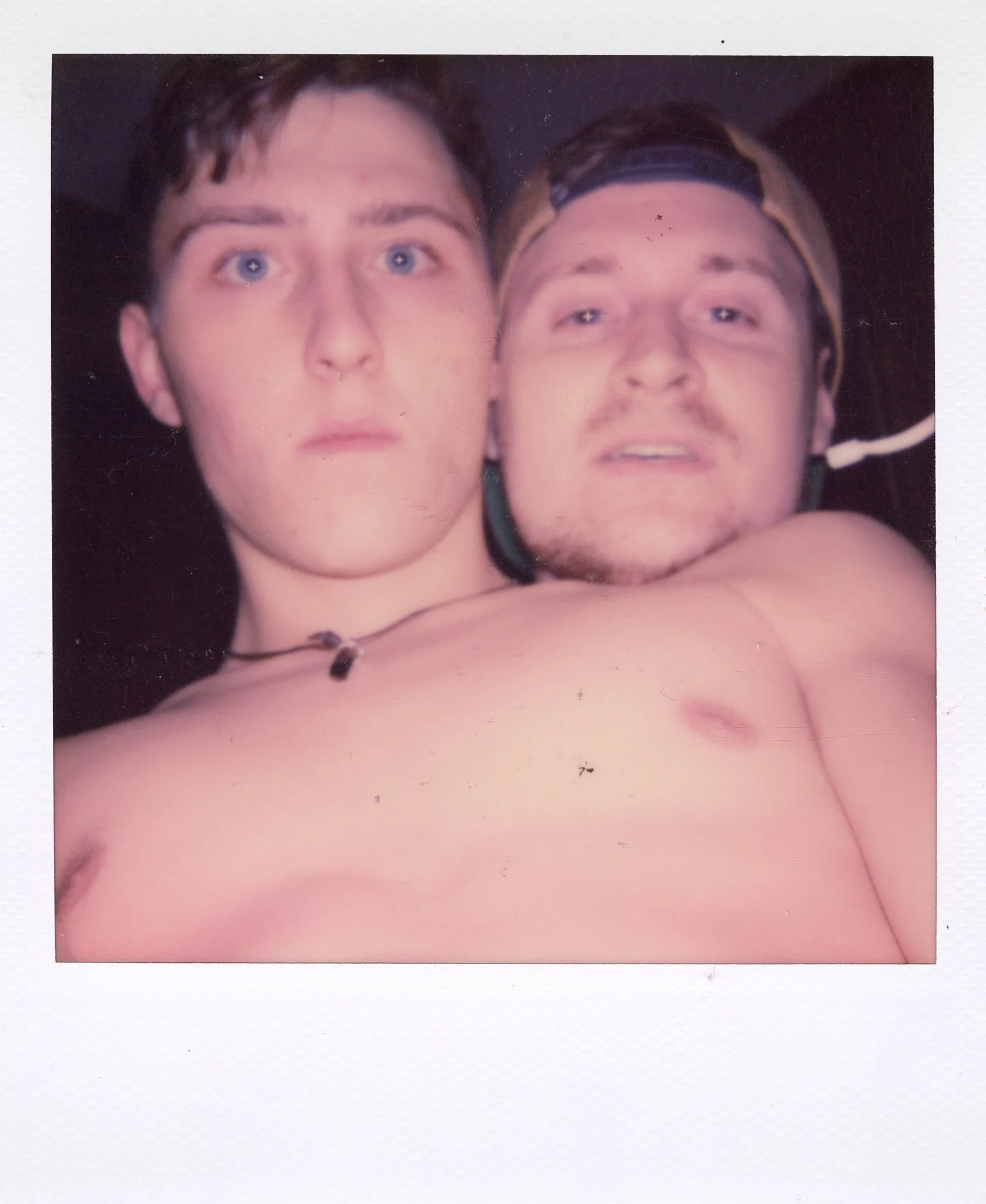 Two shirtless young men taking a close-up selfie together, one with a backward cap and the other with short hair and blue eyes.