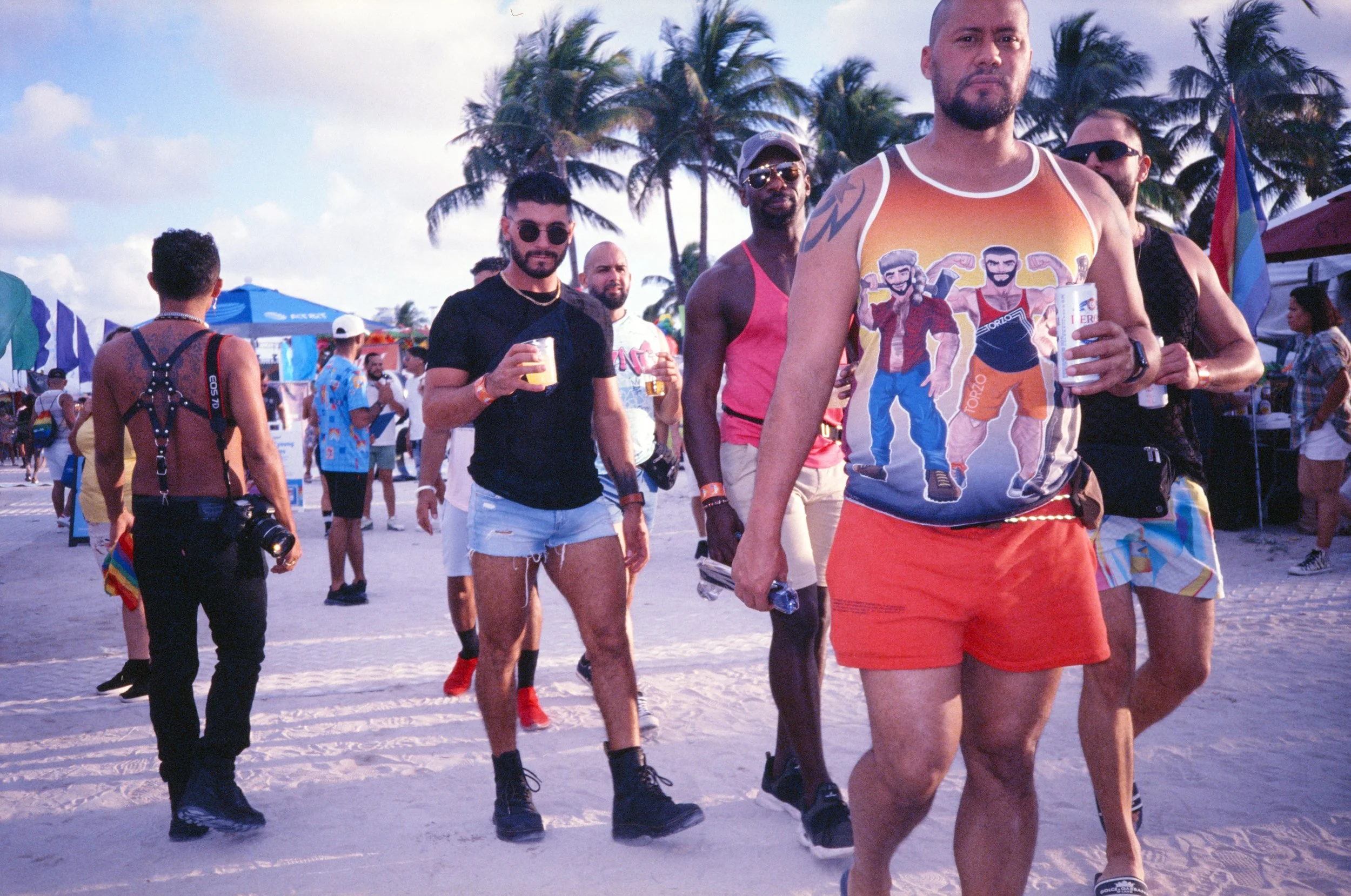 Group of men walking on a beach, holding drinks, surrounded by palm trees and beach umbrellas.