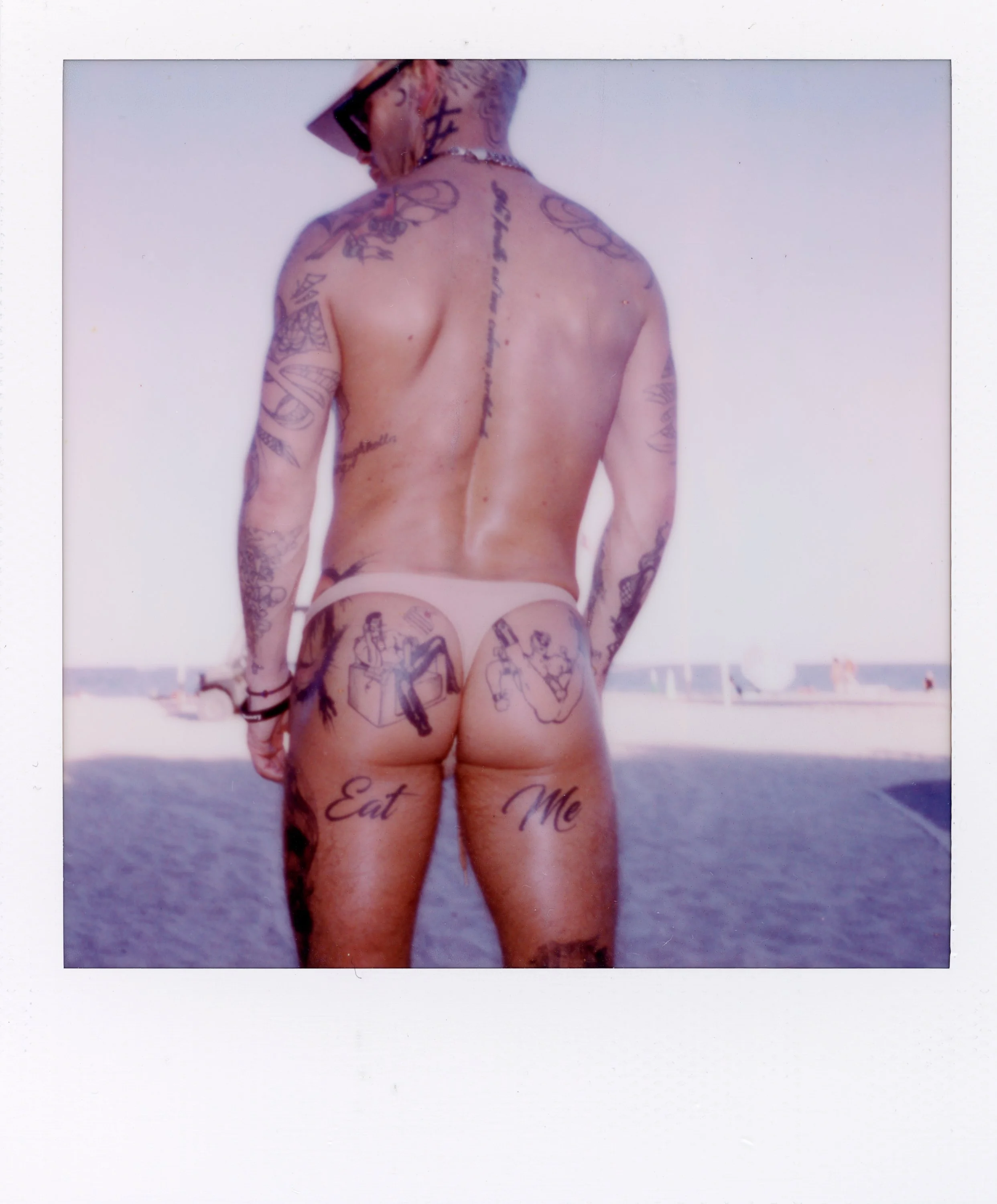 A person with tattoos standing outdoors near the water, wearing only a thong, with the words "Eat" and "Me" tattooed on their buttocks.