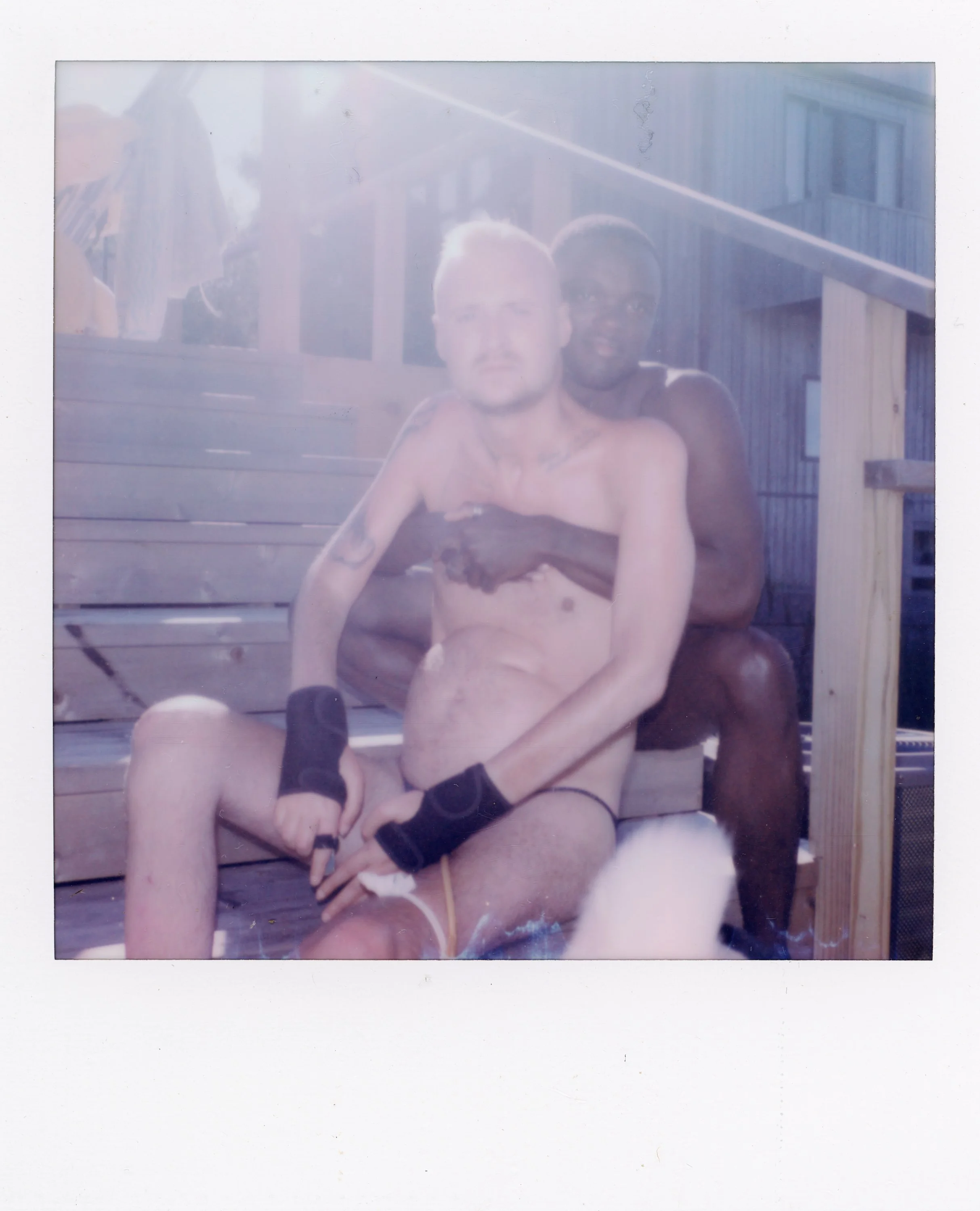 Two shirtless men sitting on wooden stairs outdoors, one with lighter skin and the other with darker skin, both wearing wristbands, with sunlight causing lens flare and overexposure in the photo.