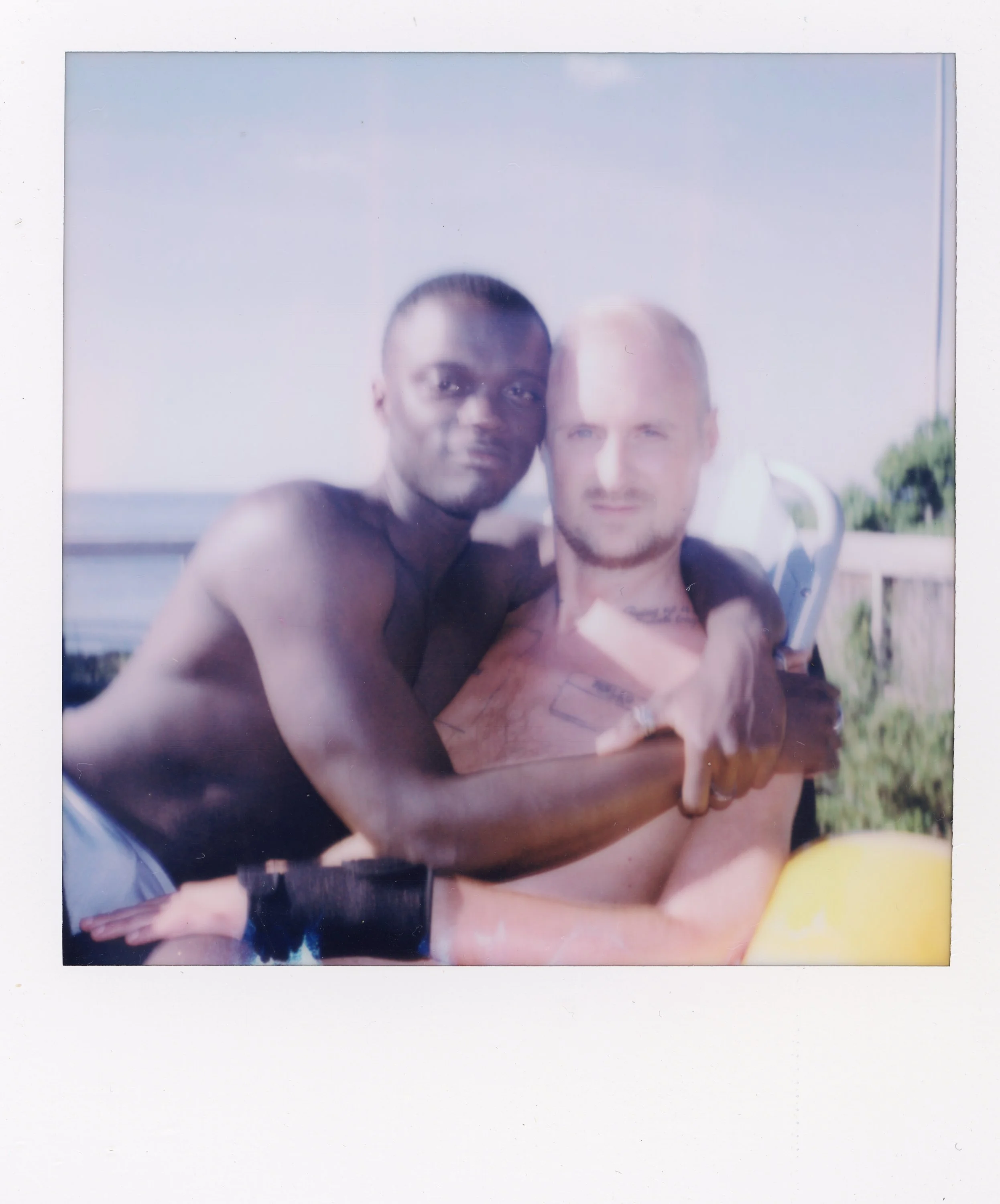 Two shirtless men embracing outdoors on a sunny day, with a railing and trees in the background.