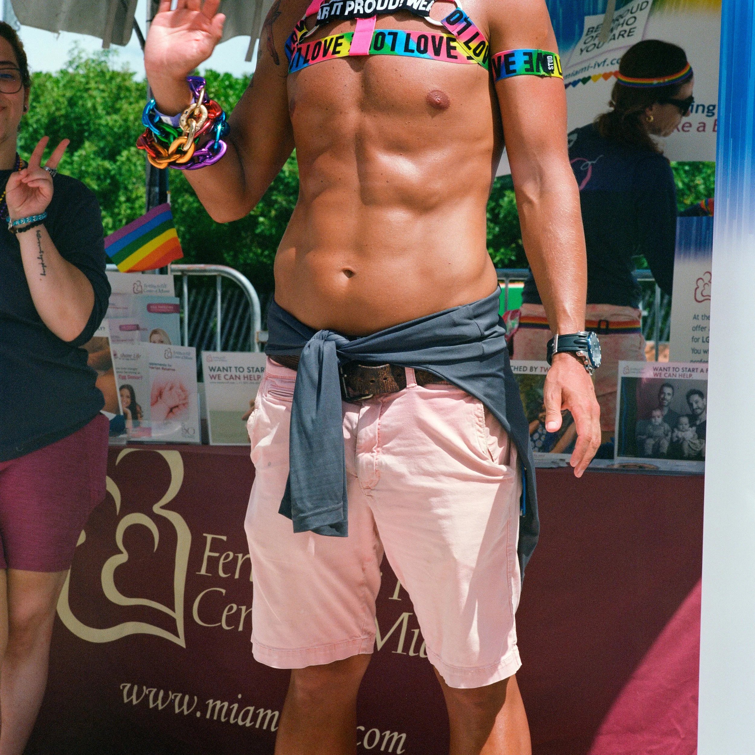 A shirtless man wearing pink shorts, a rainbow-colored 'LOVE' strap, a gray shirt tied around his waist, and colorful bracelets, standing at an outdoor event with a booth that has a rainbow flag and informational pamphlets.