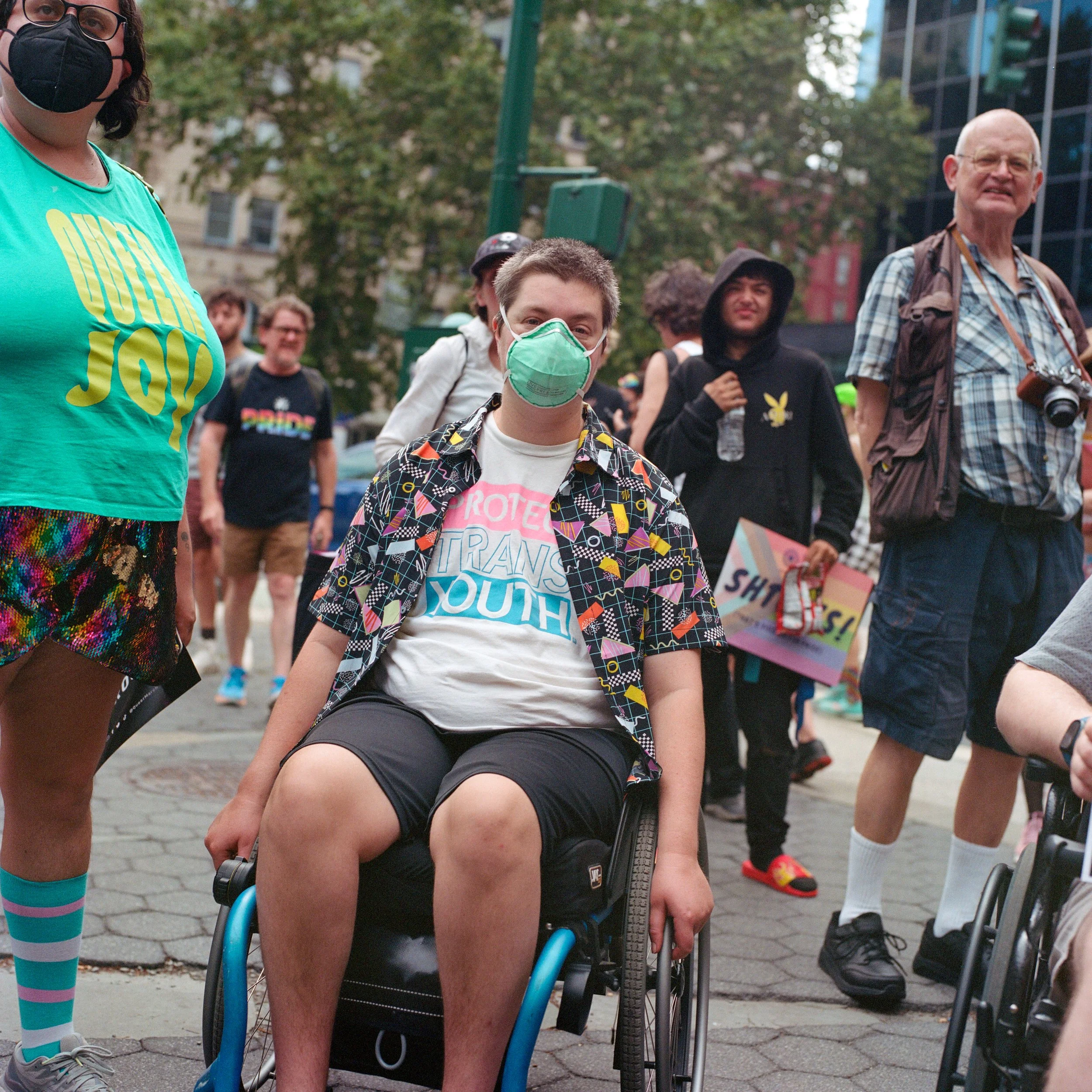 Group of people at a protest or rally, some wearing masks, outdoors in an urban area.