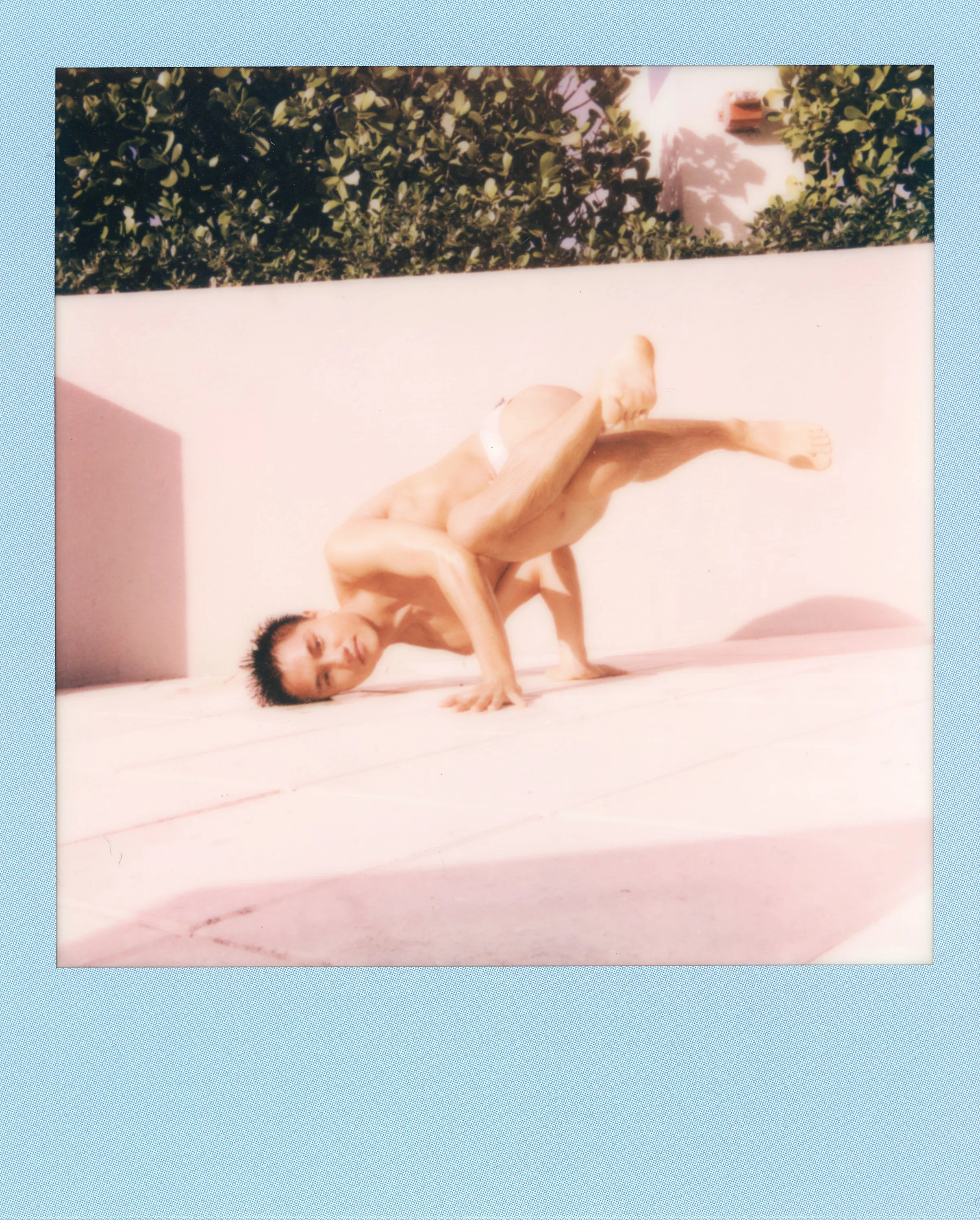 A person performing a complex pose on a pink surface outdoors, with a green leafy hedge in the background.