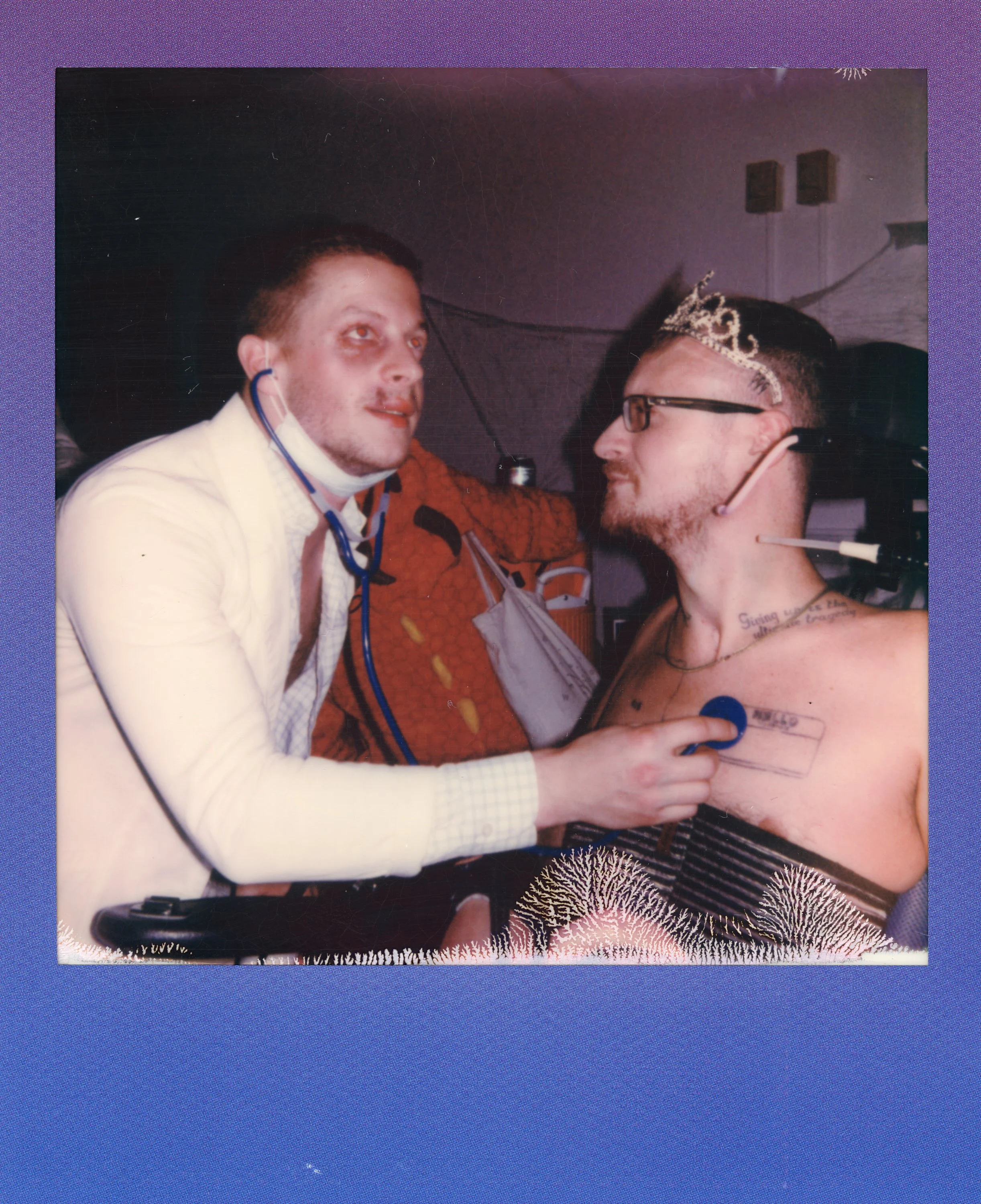 A person in a white shirt with a mask around their neck is holding a stethoscope to a shirtless man wearing glasses and a tiara, with tattoos, who is looking at them and smiling.