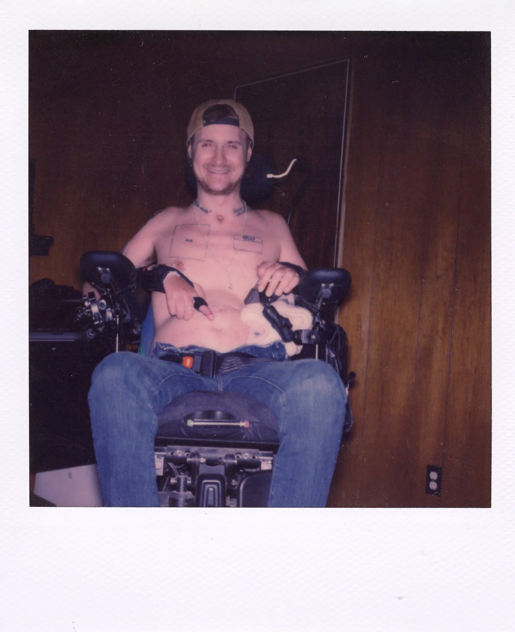 A man with long hair wearing a backward baseball cap, smiling, shirtless, sitting in a wheelchair, with tattoos on his chest and neck, indoors with wooden walls.
