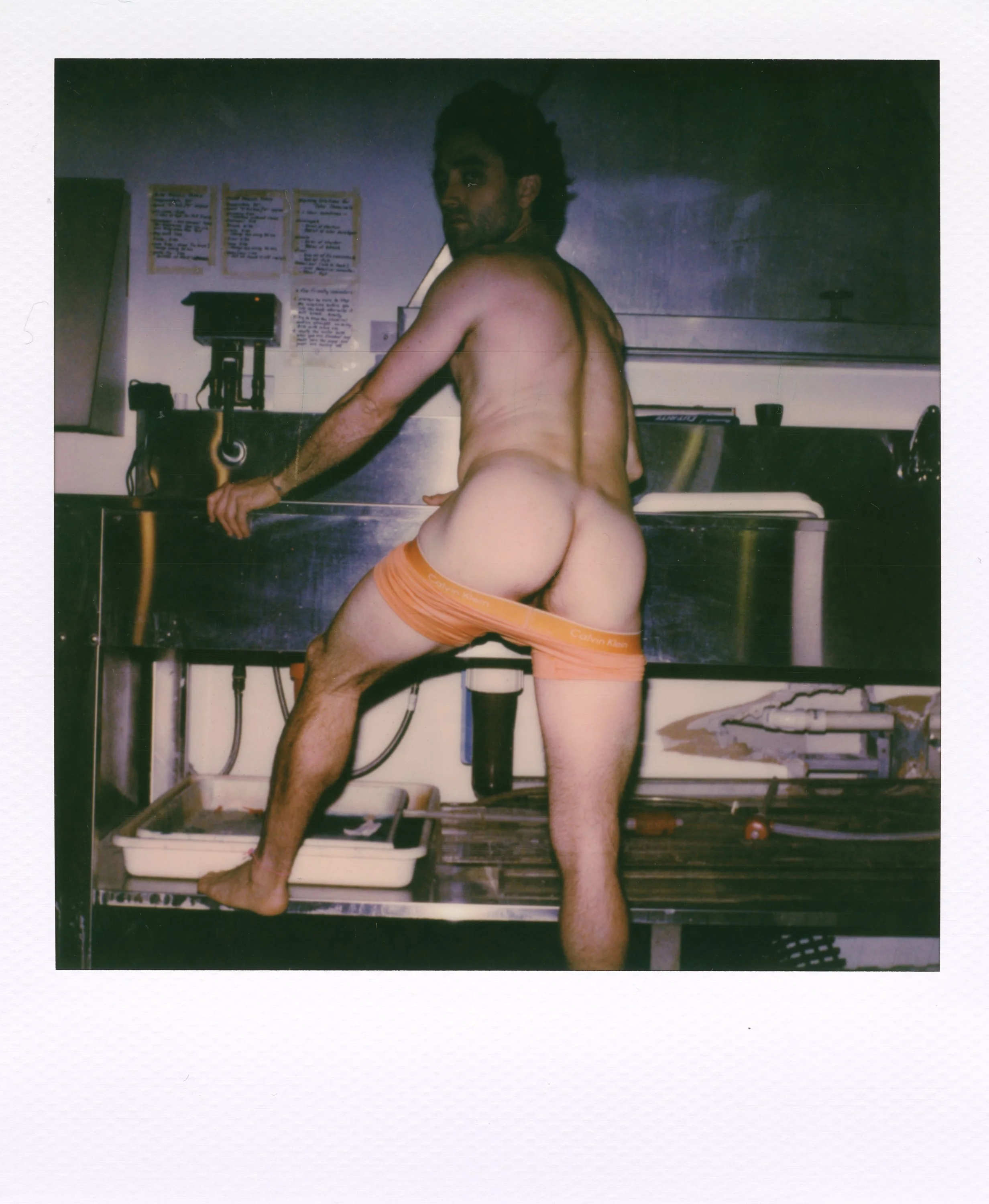 A shirtless man with curly hair and beard standing on a kitchen countertop, wearing orange Calvin Klein underwear, looking over his shoulder at the camera.