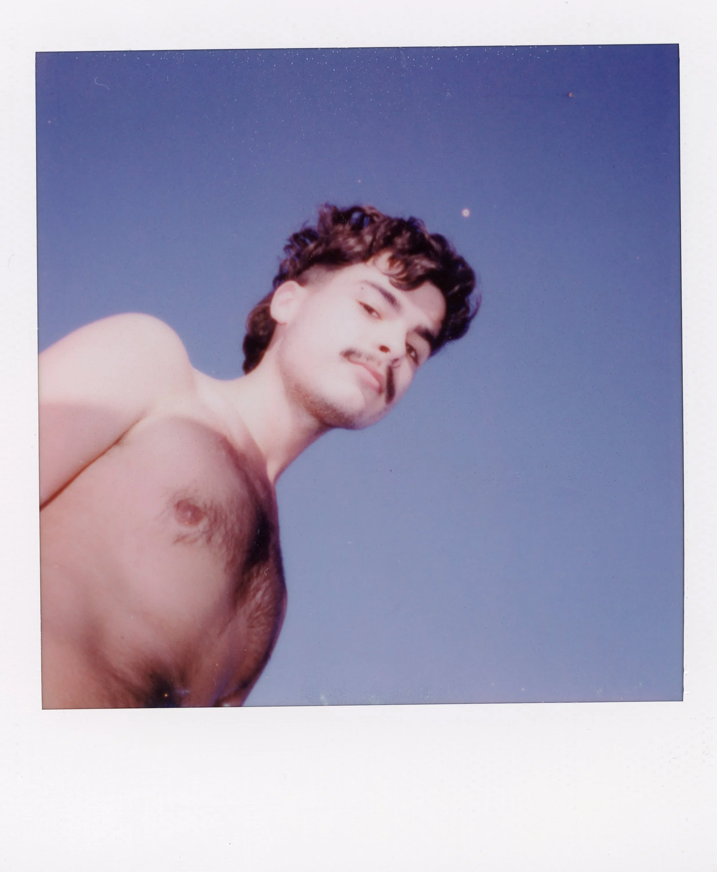 A young man with dark curly hair and a mustache, shirtless, looking down at the camera against a clear blue sky.