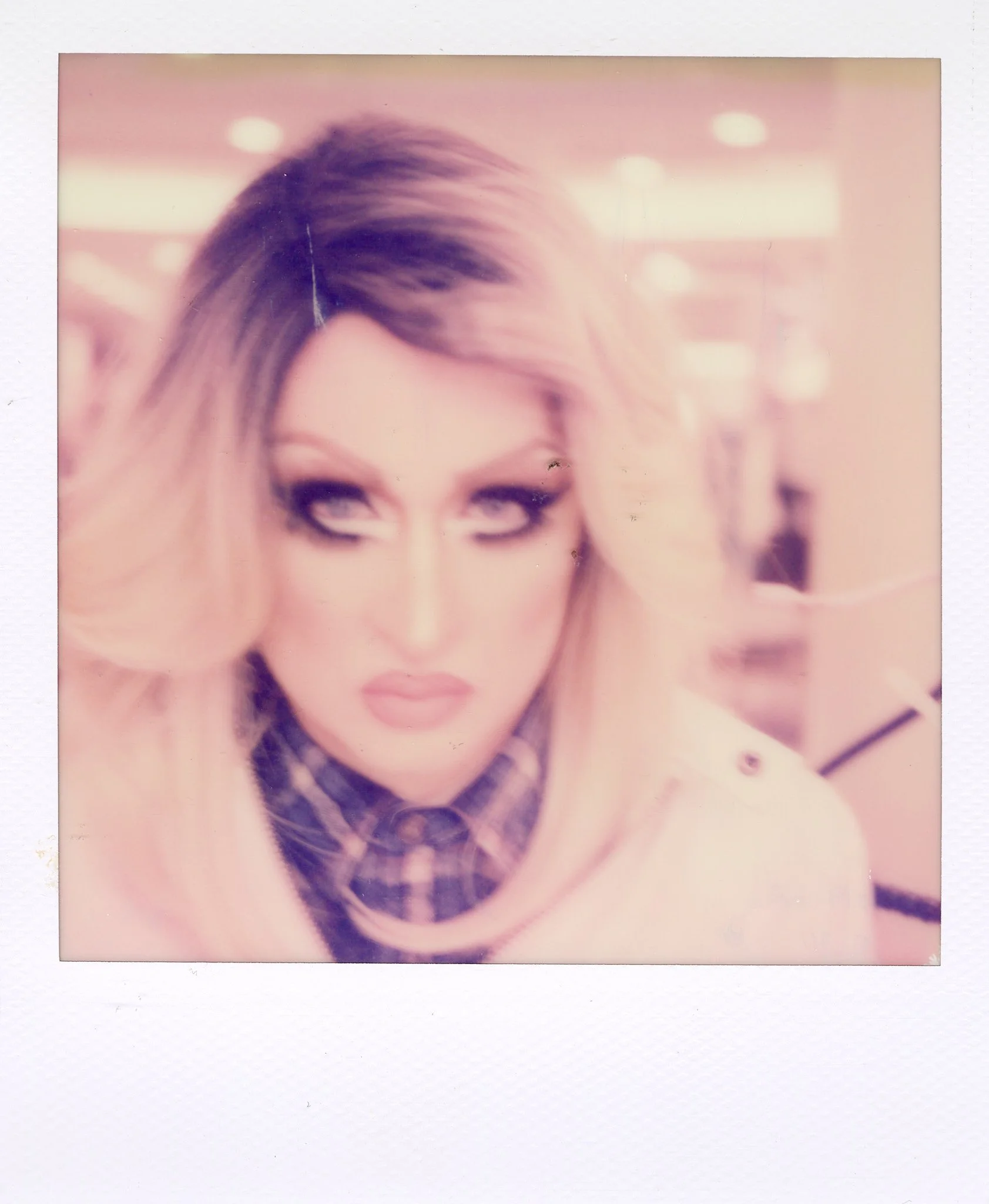 Polaroid photo of a woman with blonde hair, makeup, and a plaid shirt under a light sweater, looking to the side with an intense expression.