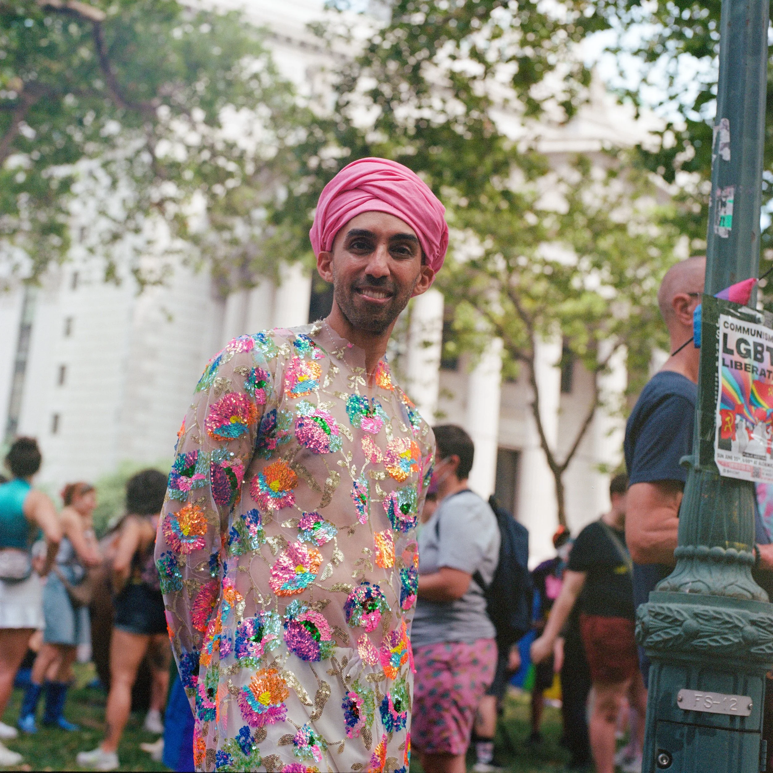 A person wearing a pink turban and a sheer, embroidered dress at a pride parade or rally, with other participants in the background.