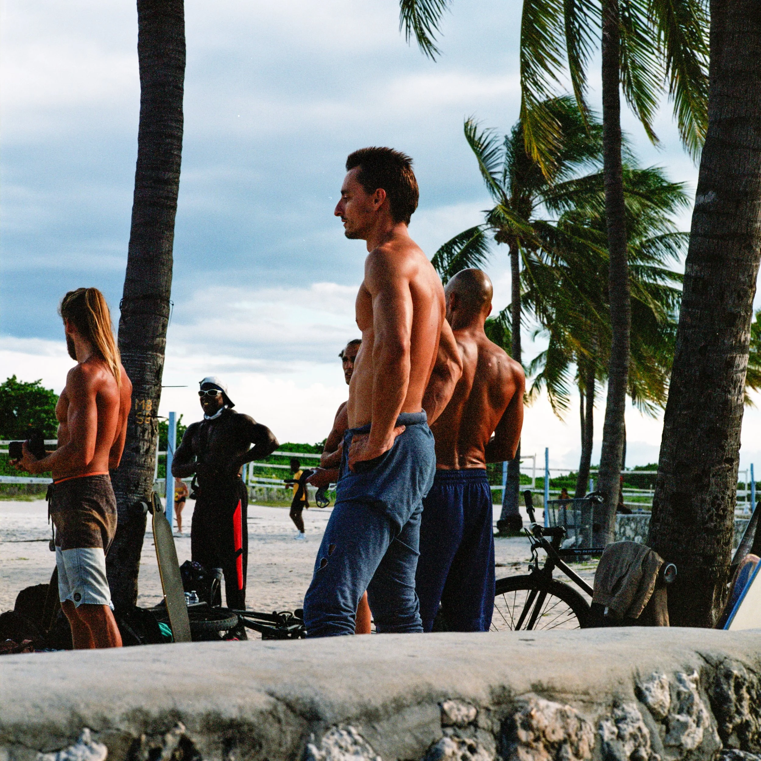 Group of shirtless men and women on a beach with palm trees, standing near bicycles and sporting equipment, during daytime.