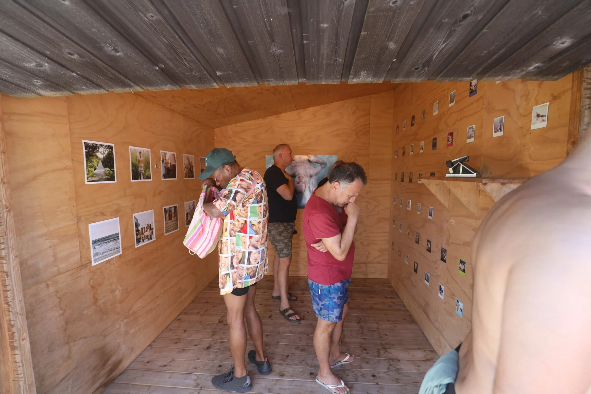 Four people in a small, wooden art gallery looking at photographs displayed on the walls, with a close-up of a person's shoulder in the foreground.
