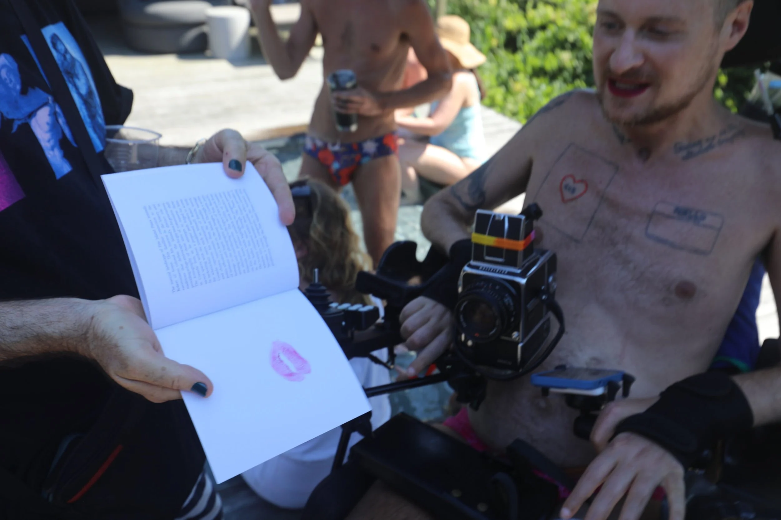 A shirtless man with tattoos receiving a kiss print on a piece of paper for a tattoo from another person. In the background, other people are relaxing outdoors, some wearing swimsuits and hats.