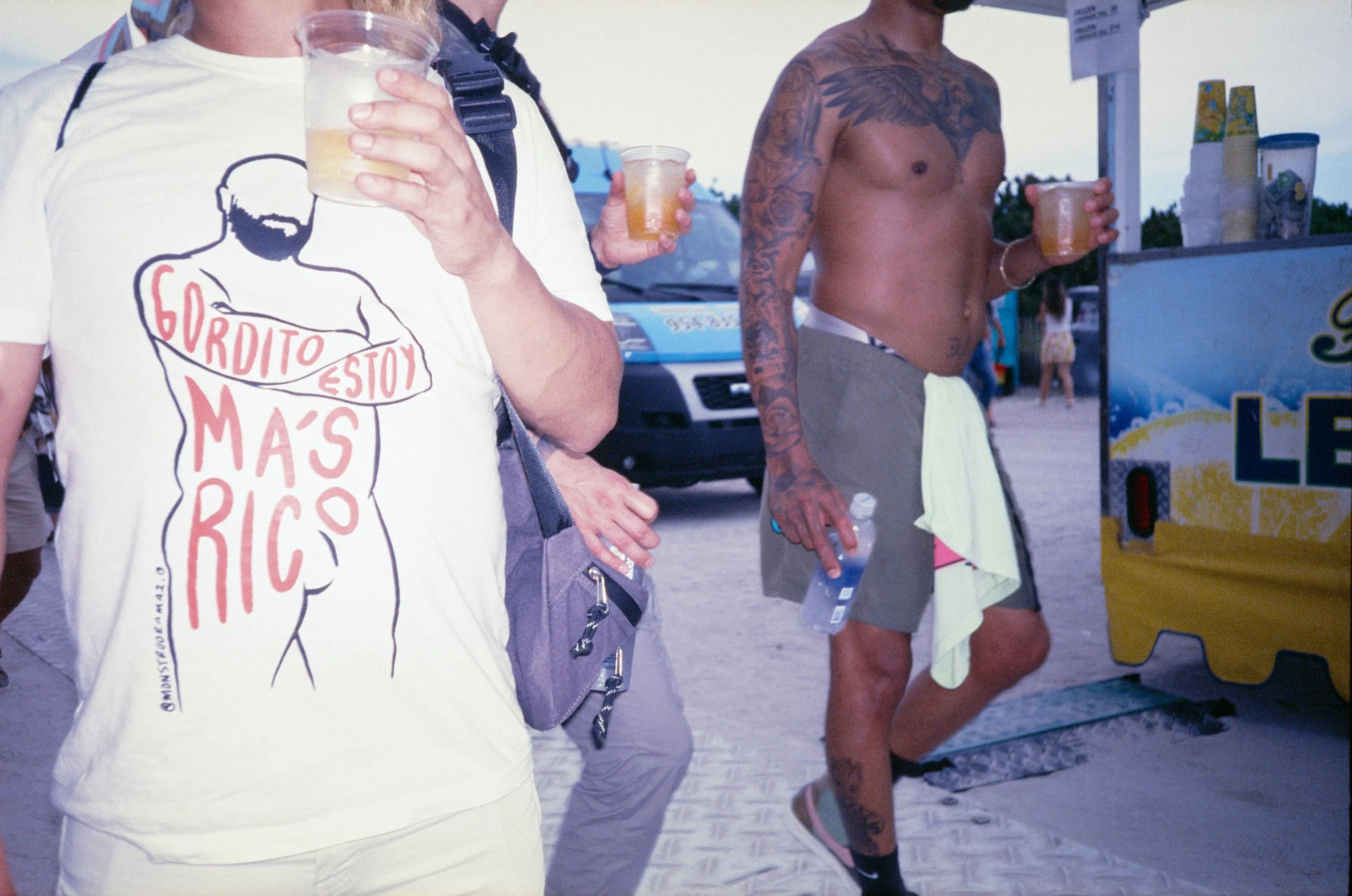 Two shirtless men with tattoos holding drinks, and a person wearing a white t-shirt with a graphic and text, at a beach or outdoor event.
