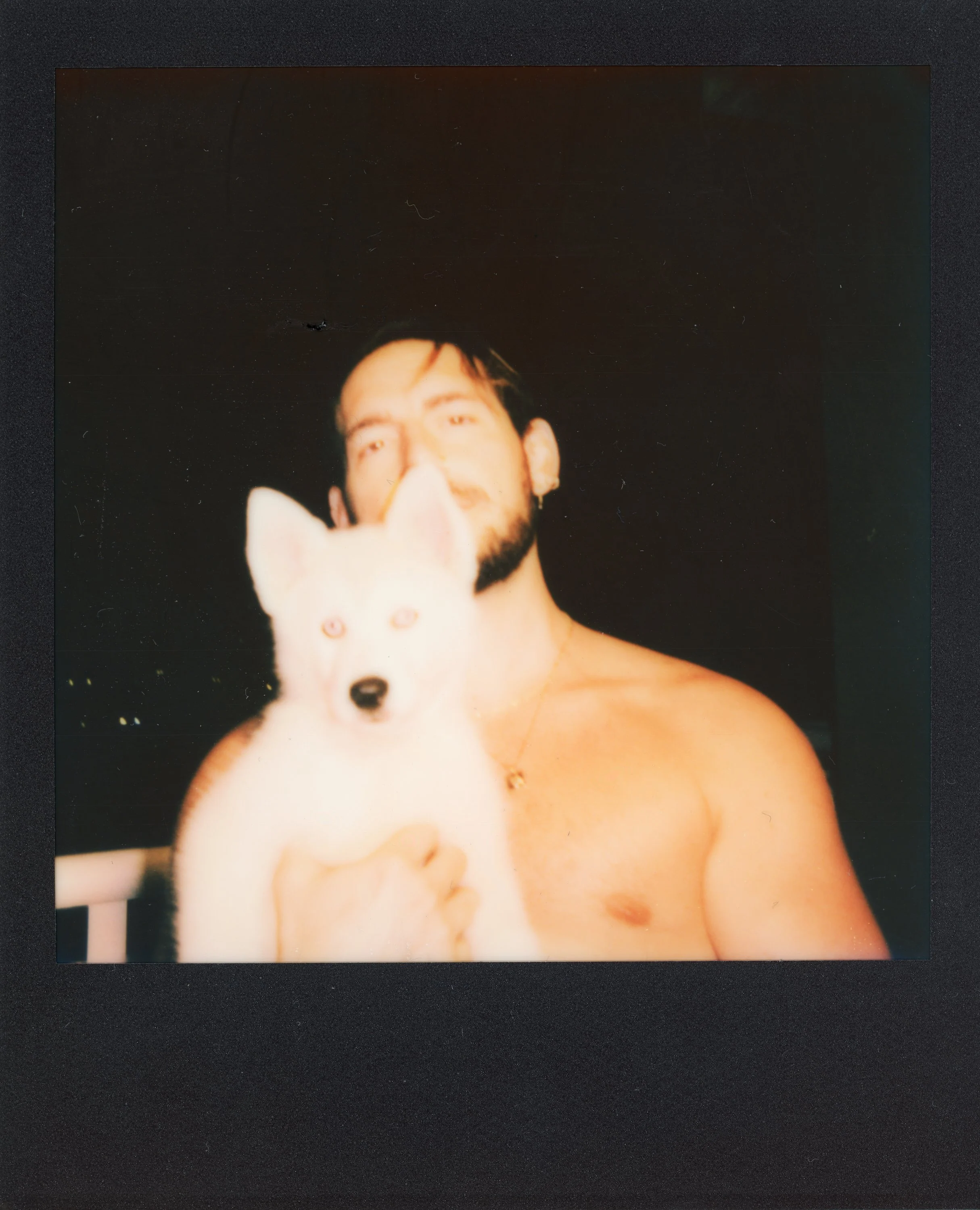 A shirtless man with dark hair and beard holding a white dog with blue eyes and upright ears, against a dark background.