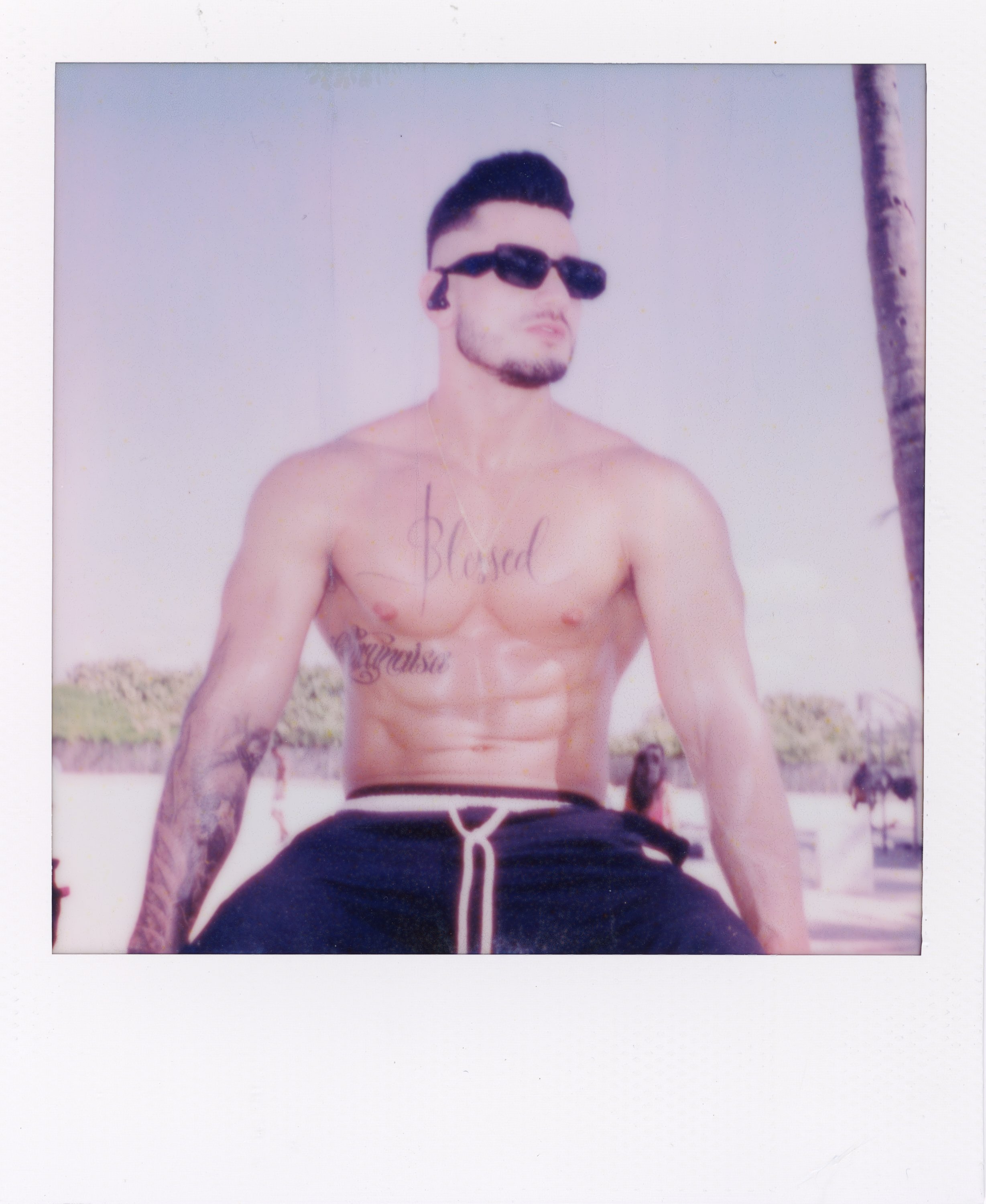 A shirtless man with tattooed arms, wearing sunglasses and black shorts, sitting outdoors near a palm tree on a sunny day.