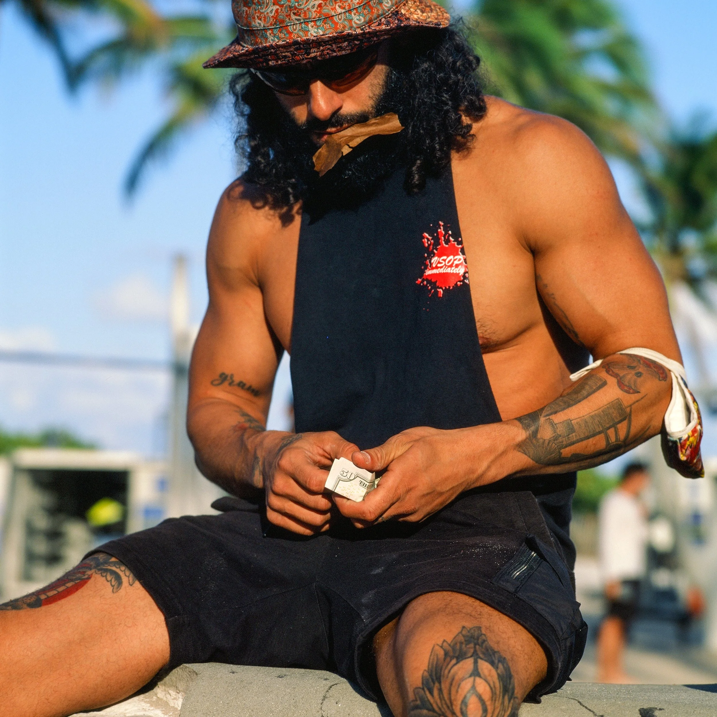 A shirtless man with tattoos, wearing sunglasses, a patterned hat, and a black sleeveless shirt, sitting outdoors with a palm tree in the background, holding money in his hands.