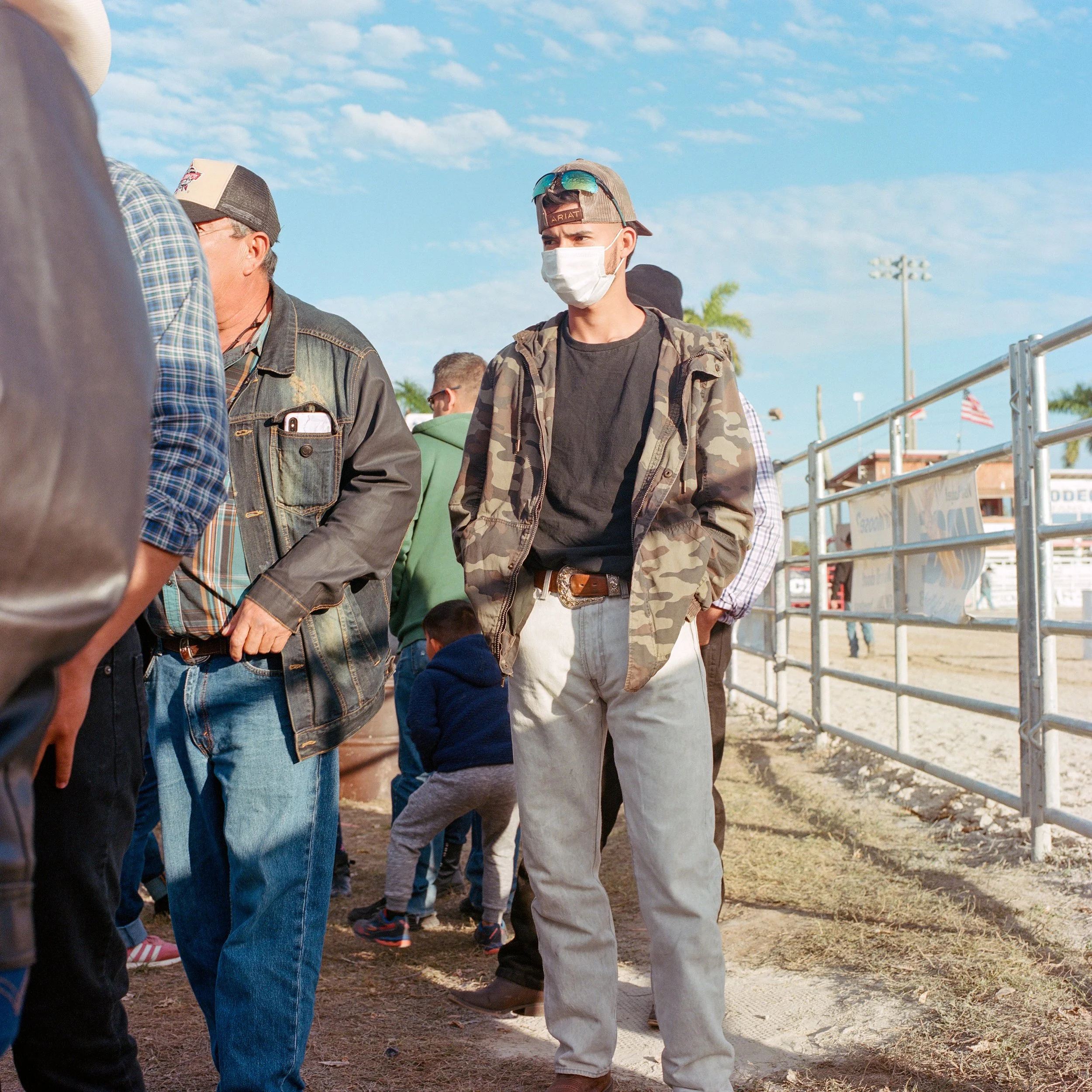 A young man wearing a face mask, camouflage jacket, black T-shirt, light-colored jeans, and sunglasses on his cap standing outdoors near a fenced area with other people around.