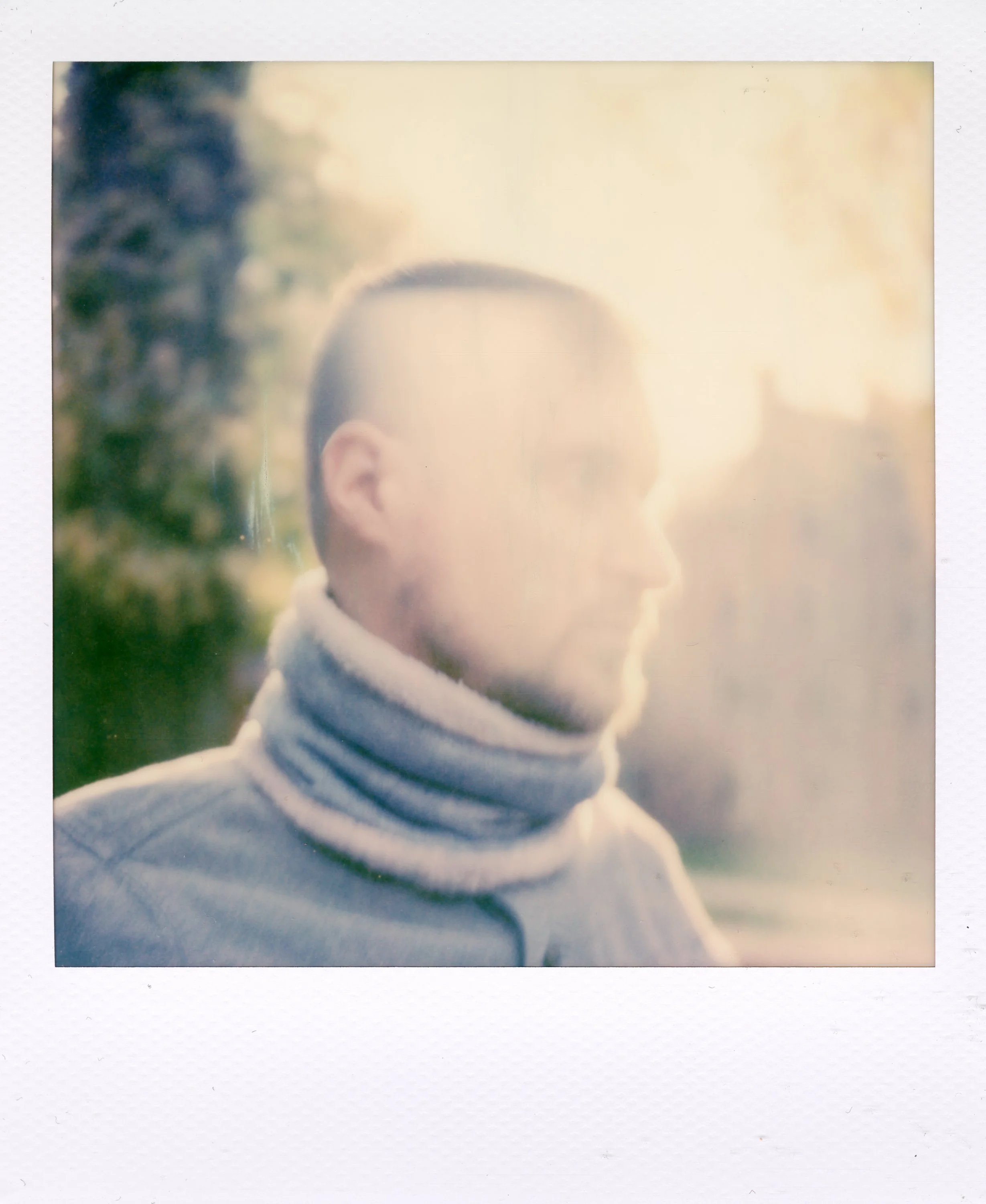 Polaroid photo of a man with his eyes closed, wearing a gray zip-up jacket and a striped scarf around his neck, illuminated by bright sunlight, with blurred trees in the background.