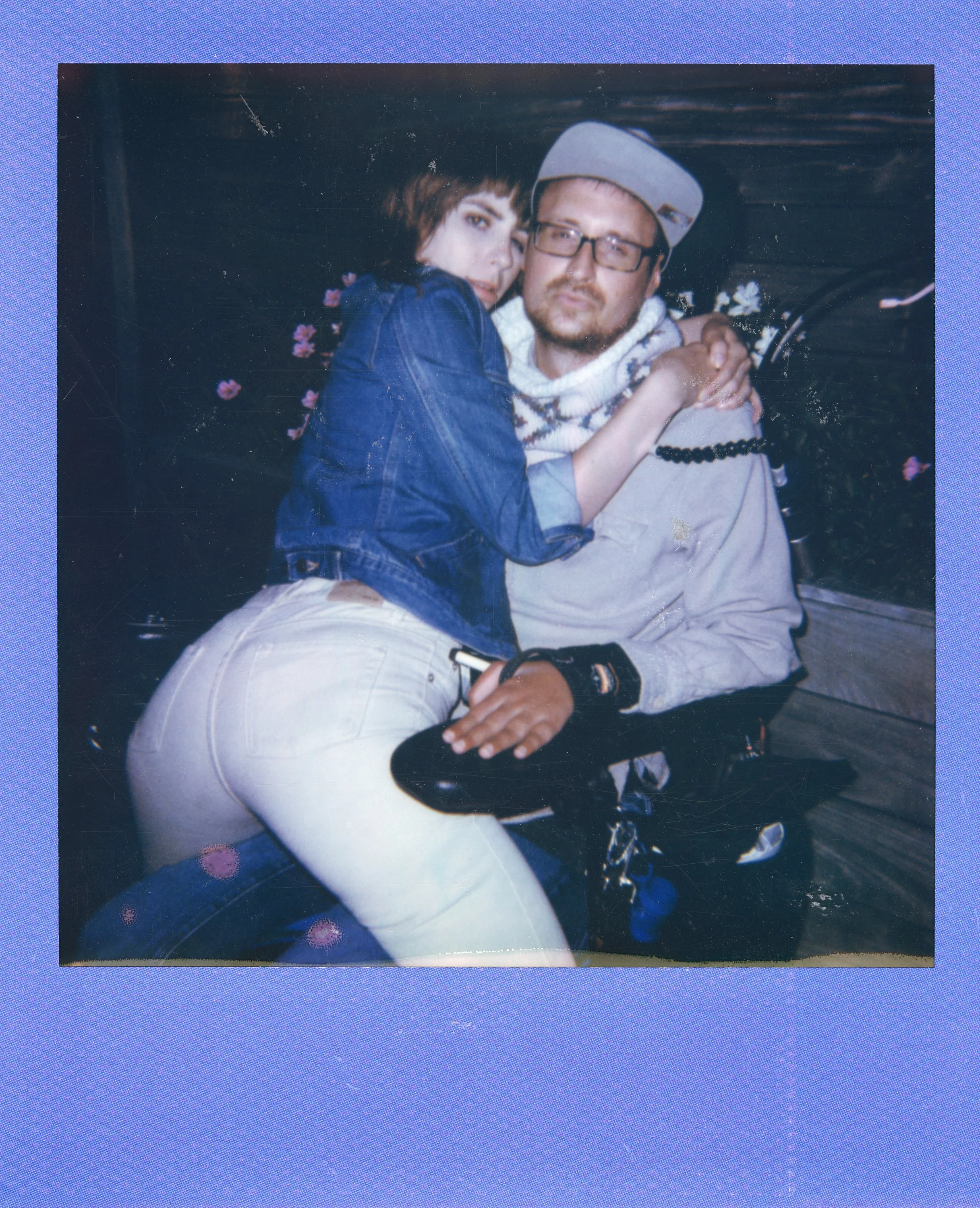 Young woman with short dark hair wearing a denim jacket, sitting on a man's lap, with her arms around his neck, in front of a backdrop with pink flowers. The man has glasses, a cap, and a sweater, and is seated on a bench or similar surface.