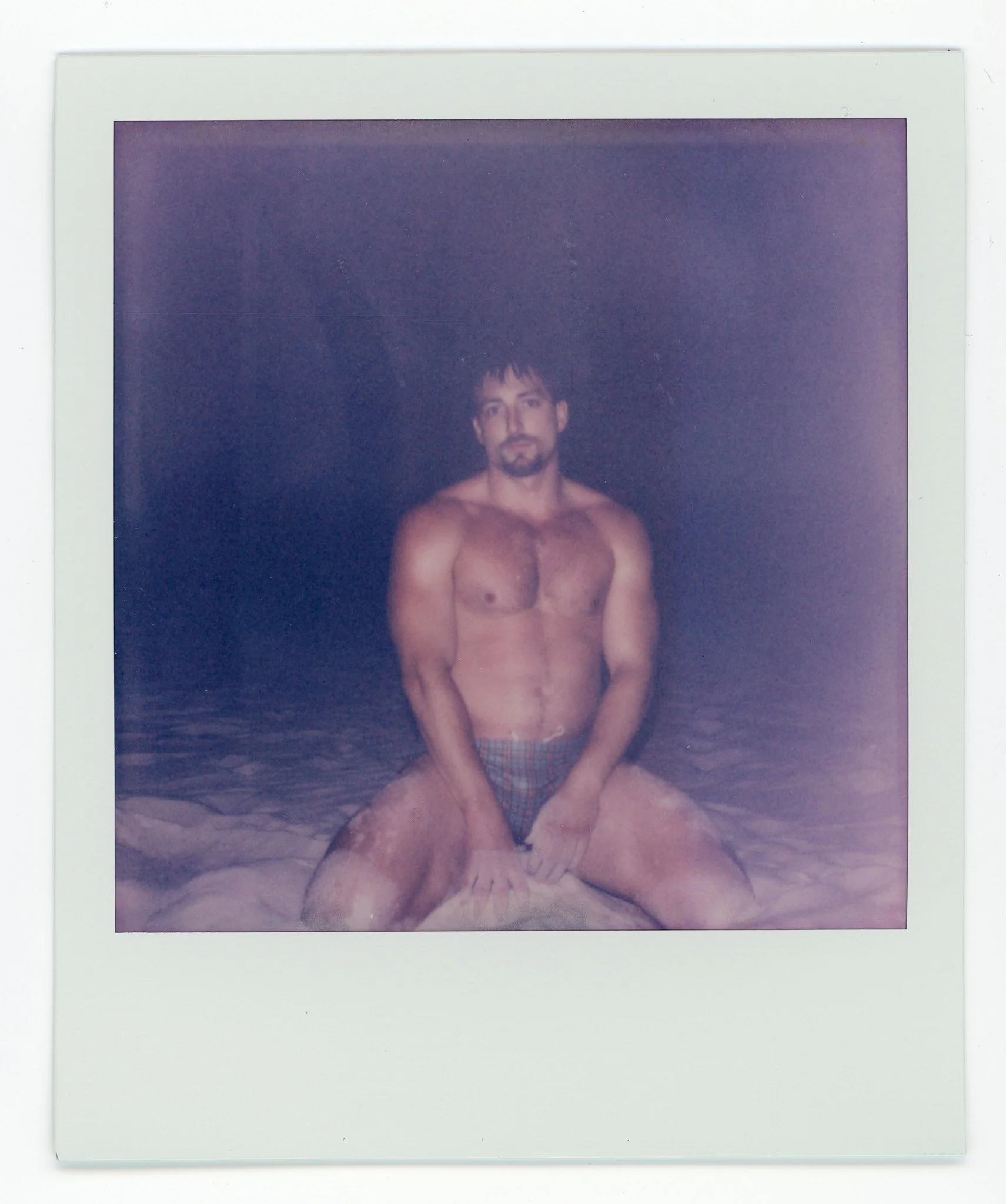 A shirtless man with dark hair and beard sitting on the ground with his legs spread apart, in front of a dark background.