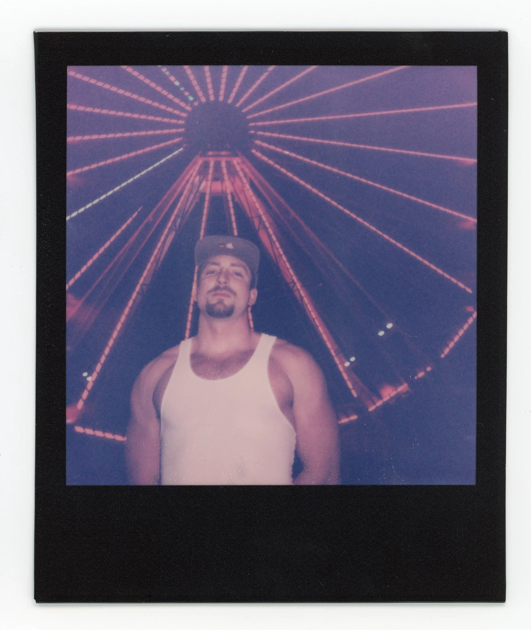 A man in a white tank top and baseball cap standing in front of a lit Ferris wheel at night.