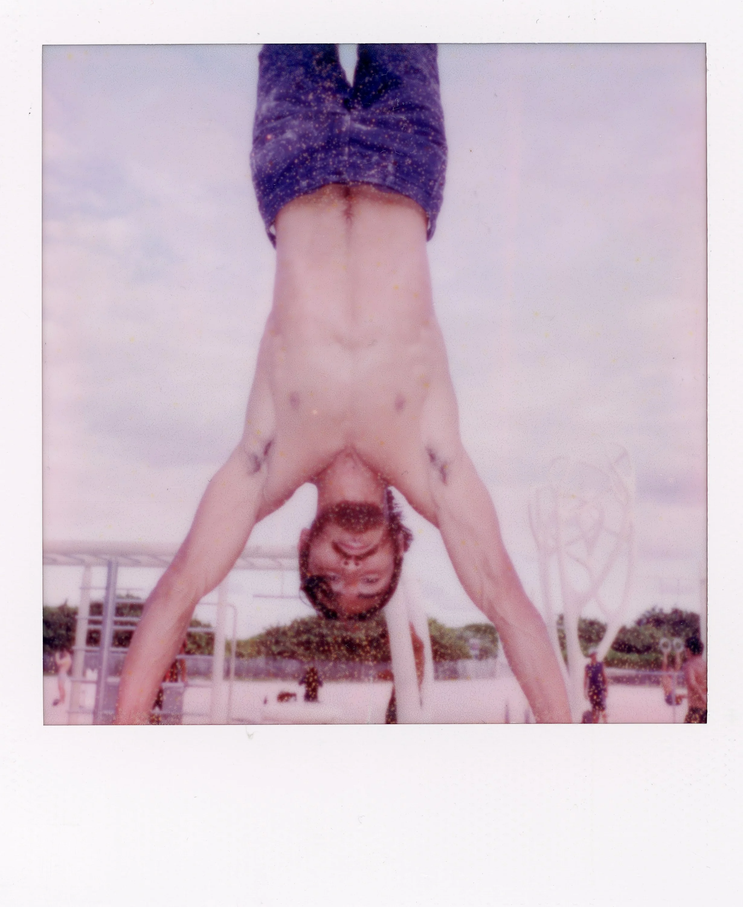 A smiling man is performing a handstand outdoors at a skatepark, with a partly cloudy sky in the background.