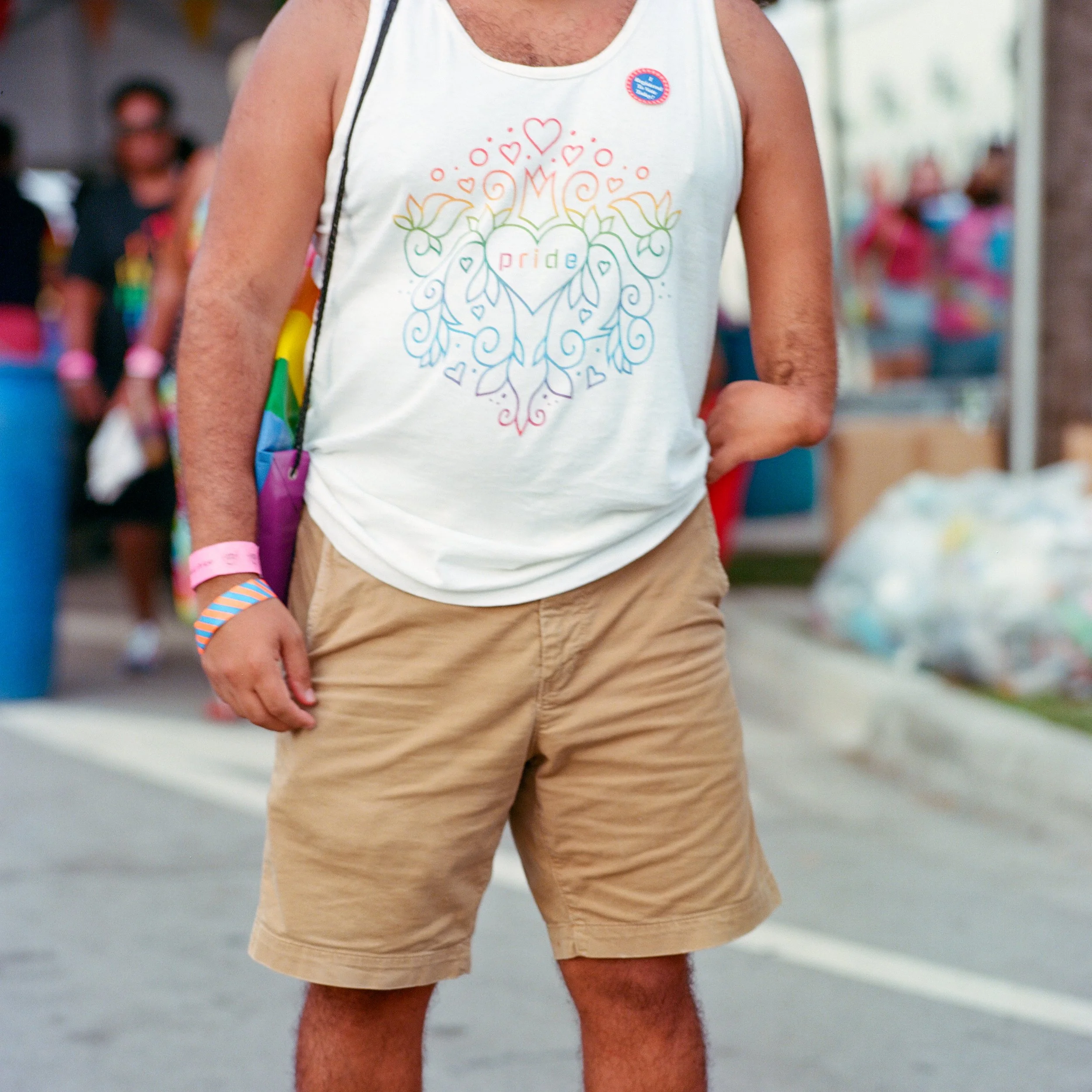 Close-up of a person wearing a white sleeveless shirt with rainbow-colored pride-themed design and the word 'pride' written on it, beige shorts, and a rainbow wristband at a pride event.